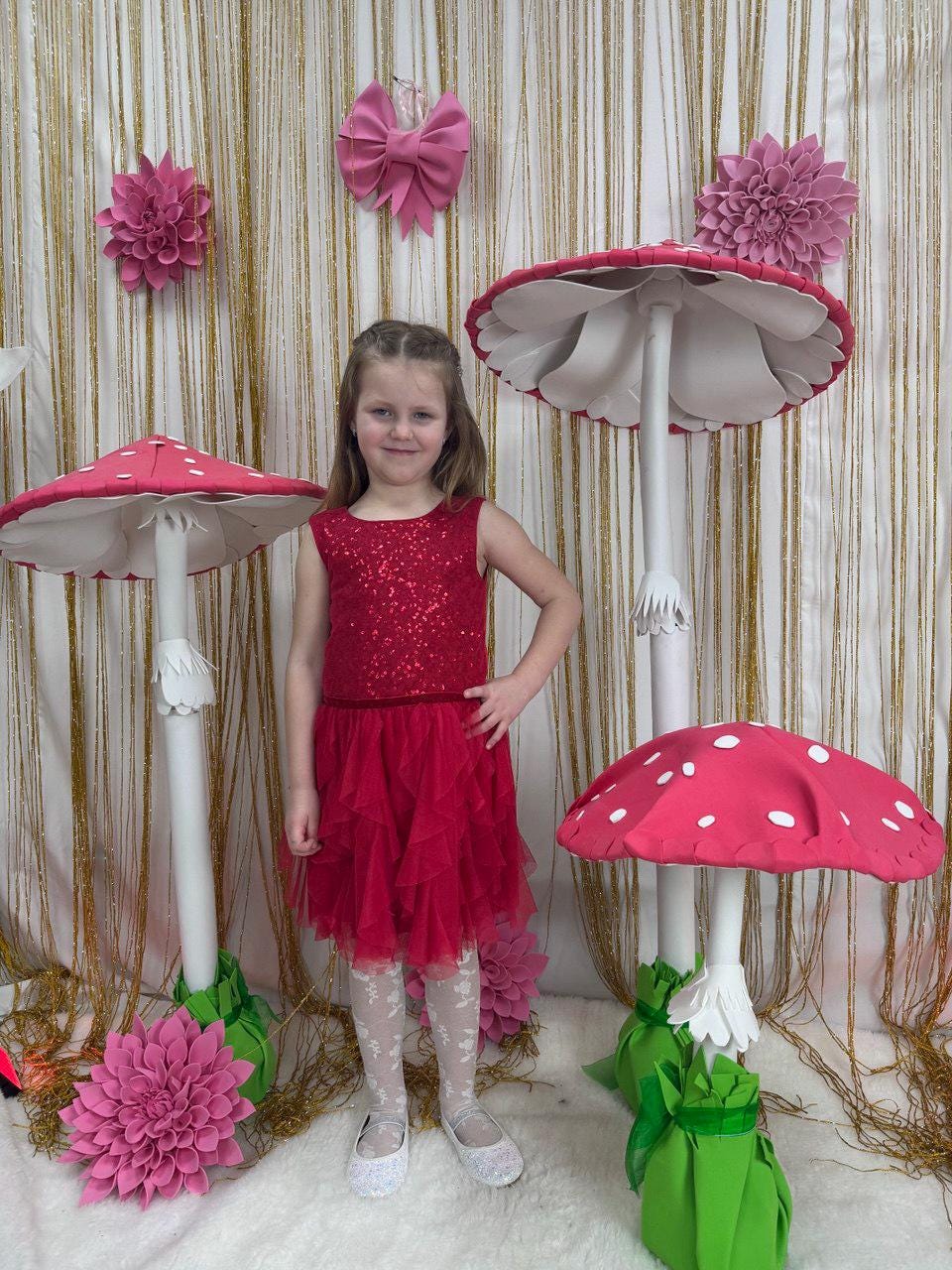 May include: A young girl in a red sequined dress and white tights stands in front of large, whimsical mushroom decorations. The mushrooms have red caps with white spots and white stems. Pink floral decorations and a pink bow adorn the background.