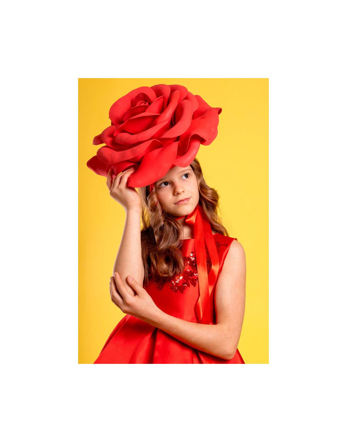 May include: A young person wearing a vibrant red dress and a large, oversized red rose headpiece. The dress features a satin finish and a ribbon detail. The background is a bright yellow, creating a striking contrast with the red attire.