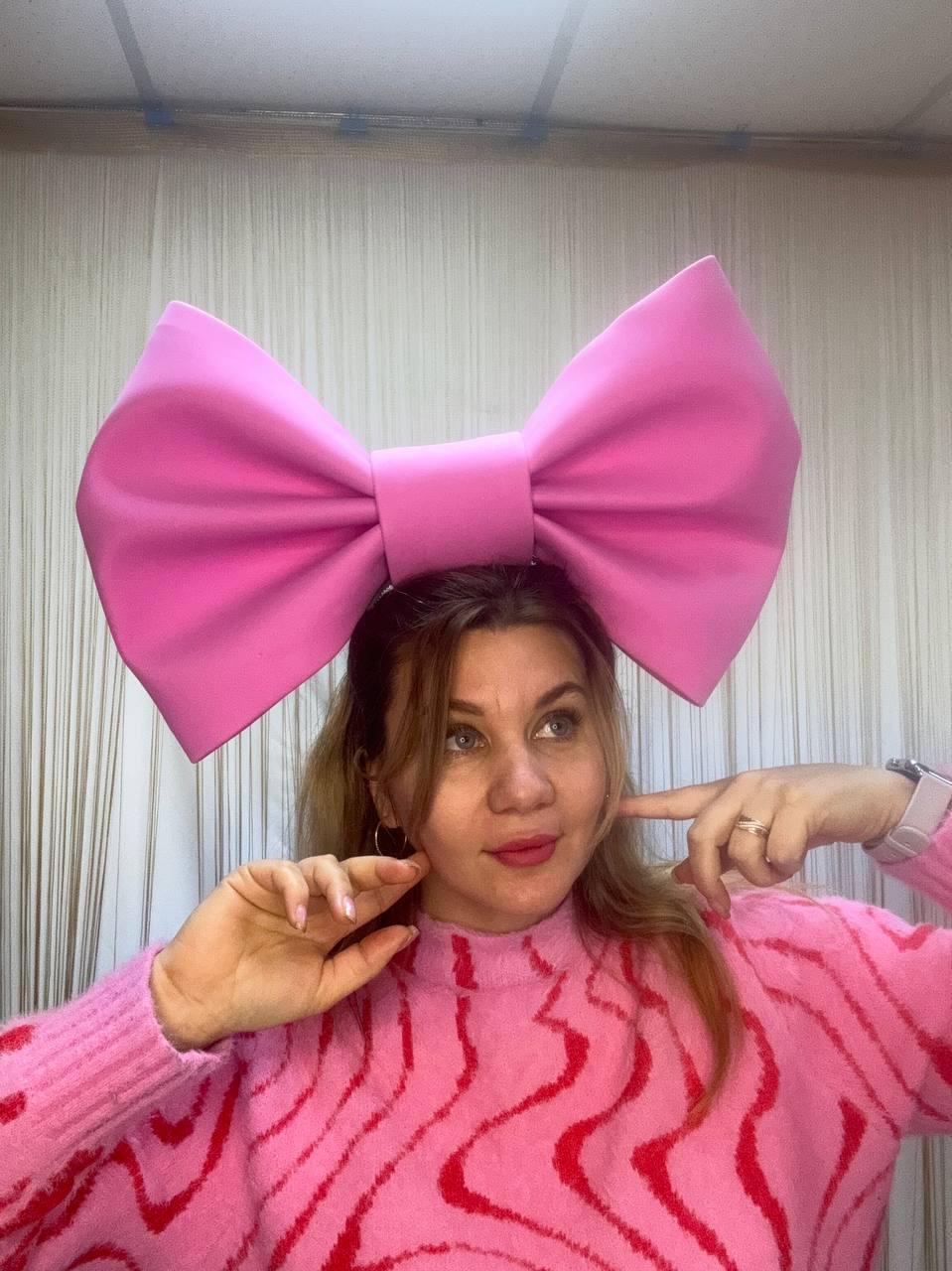 May include: A large, bright pink bow headband. The bow is the focal point, with a symmetrical design. The person in the image is wearing a pink sweater with a red wavy pattern.
