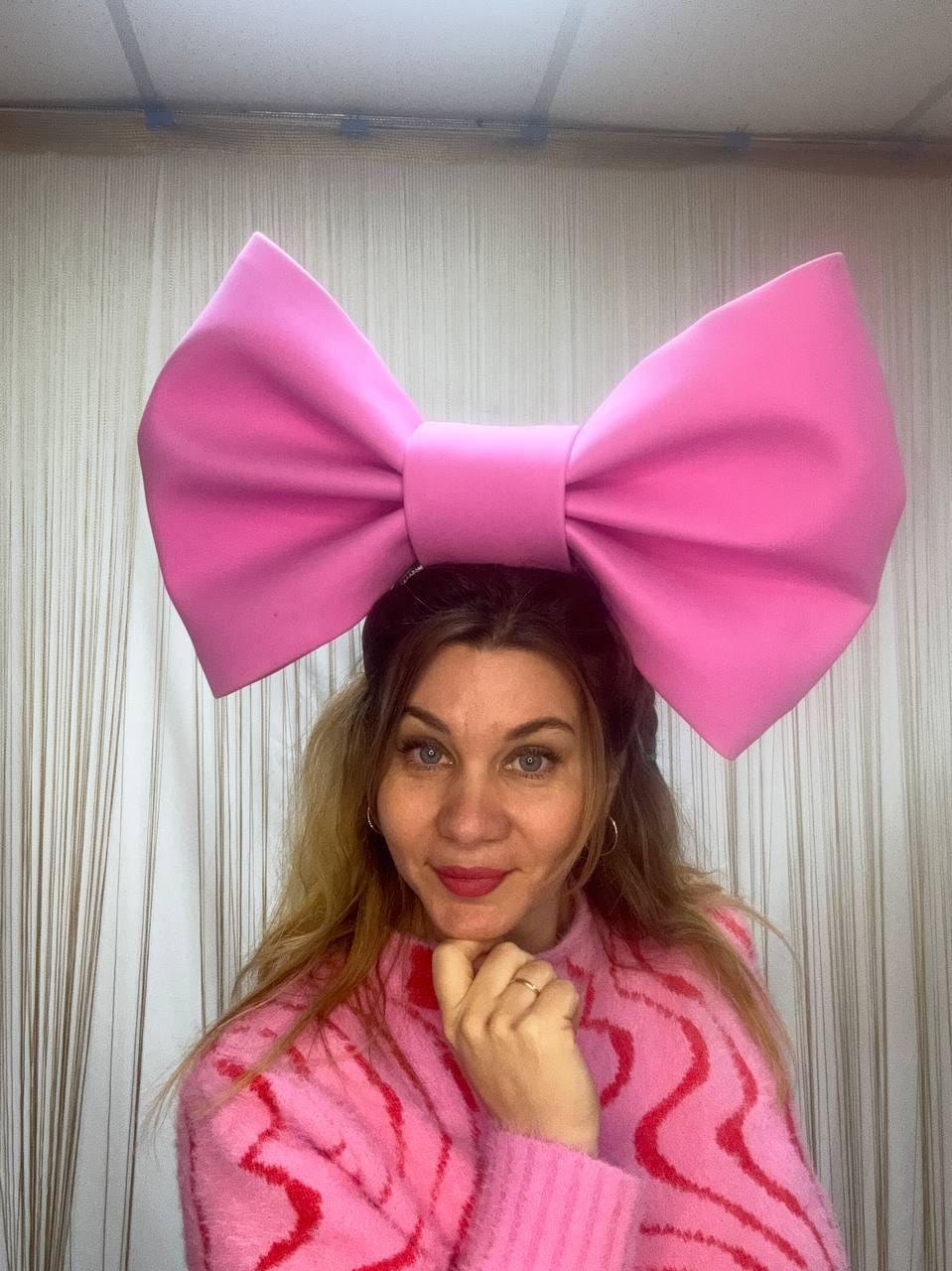 May include: A large, bright pink bow headband. The bow is oversized and made of a solid material. The person in the image is wearing a pink sweater with a red wavy pattern.
