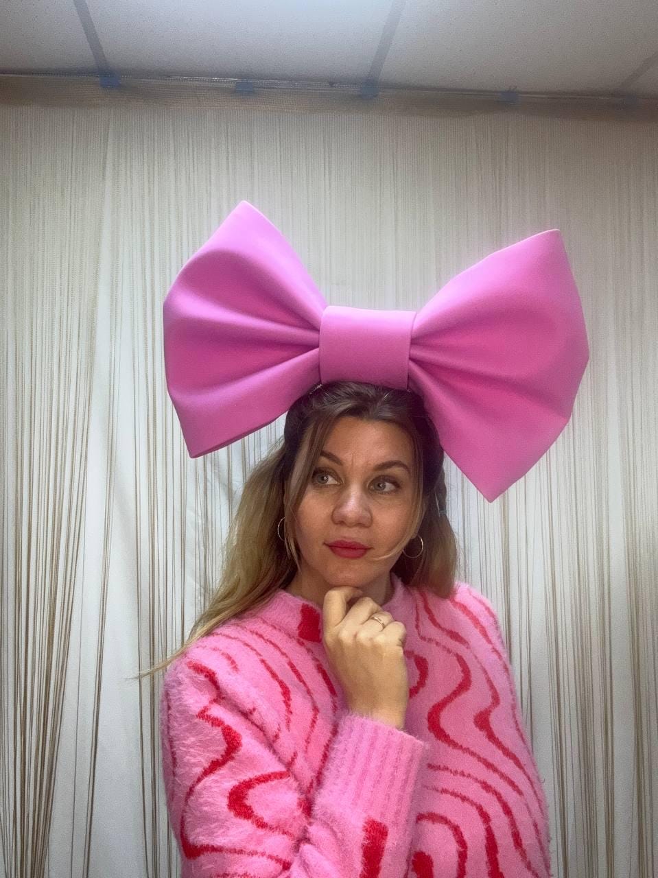 May include: A large, bright pink bow headband. The bow is oversized and the same shade of pink as the sweater. The sweater has a pink and red wavy pattern. The background is a neutral color.