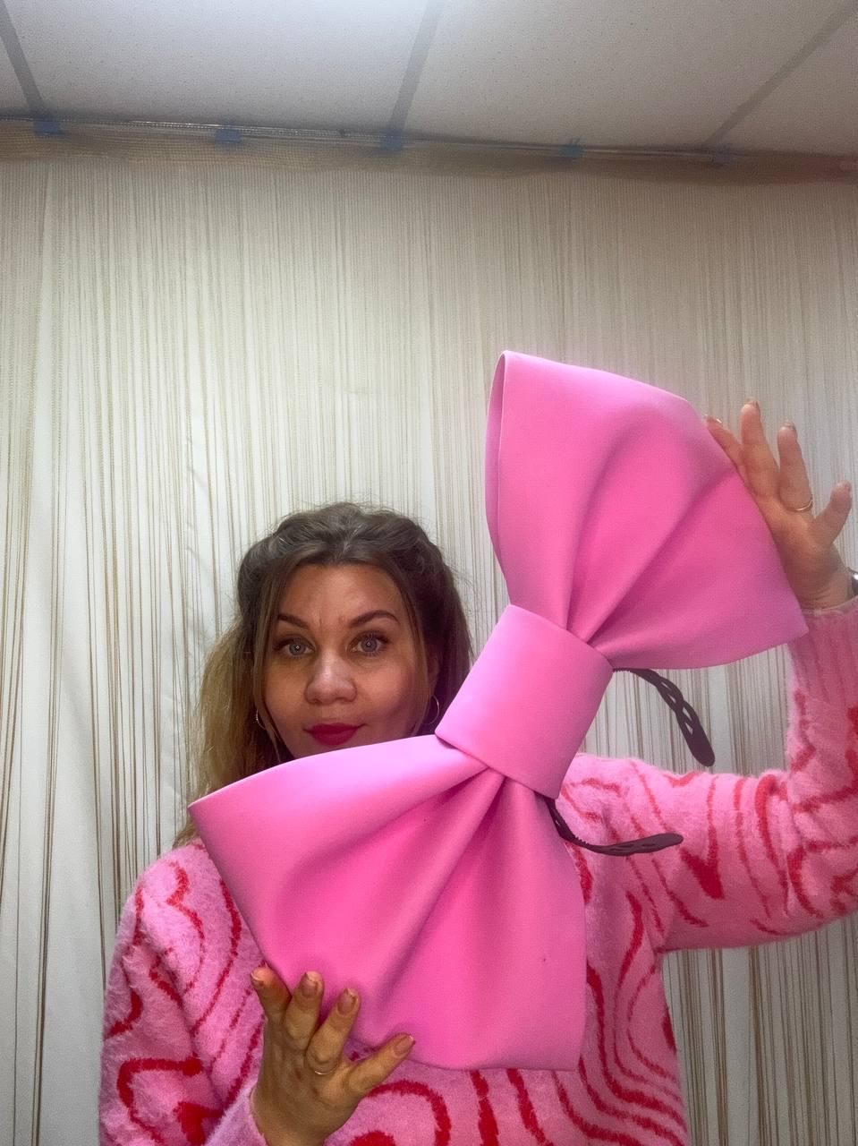 May include: A large, bright pink bow-shaped handbag. The bag is held up in front of a person wearing a pink sweater. The bow has a thick, rounded shape with a central band and a black strap.