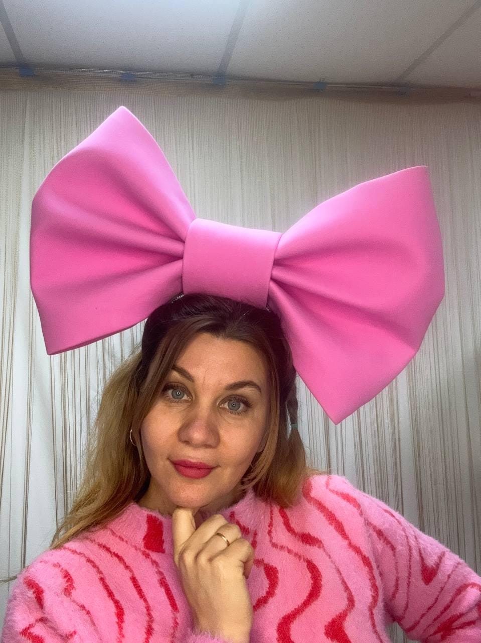 May include: A large, bright pink bow headband. The bow is oversized and the focal point of the image. The person in the image is wearing a pink sweater with a wavy pattern. The bow is a bold fashion statement.