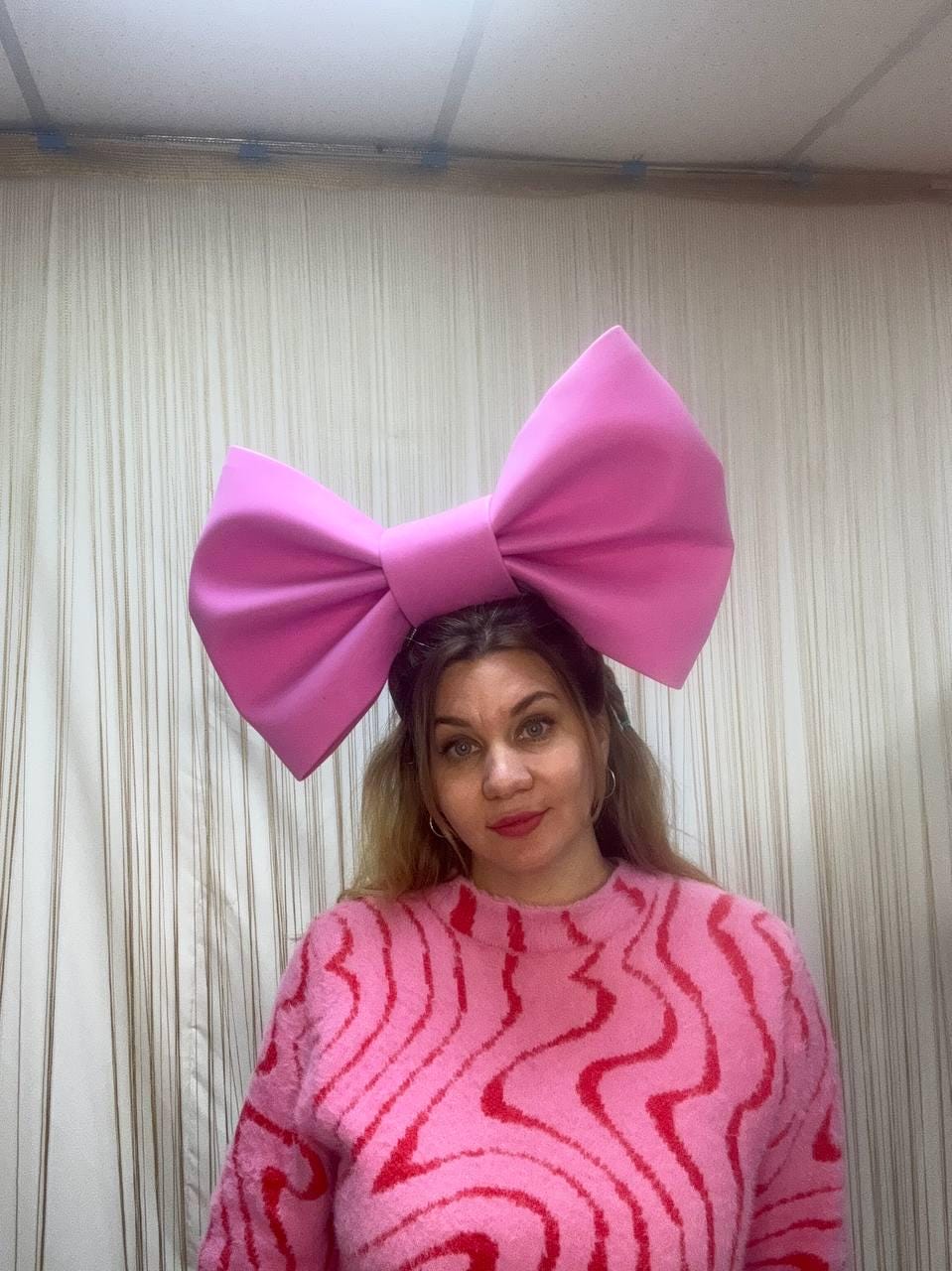 May include: A large, bright pink bow headband. The bow is the focal point, with a voluminous design. The person in the image is wearing a pink sweater with a red wavy pattern. The background is a neutral-toned curtain.