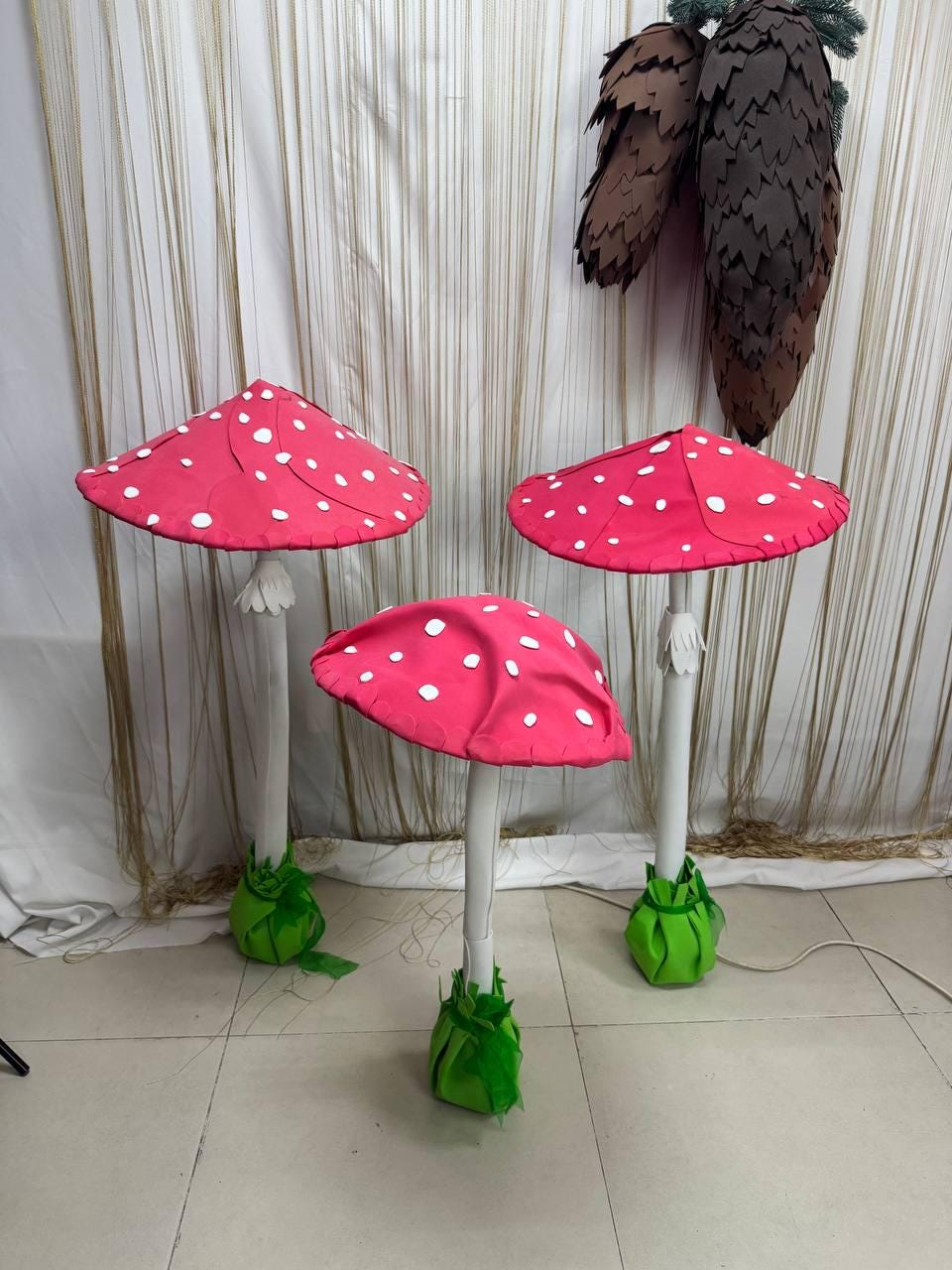 May include: Three whimsical mushroom decorations. Each mushroom has a bright red cap with white polka dots, a white stem, and a green base. The mushrooms are arranged in a row, with a backdrop of a curtain.