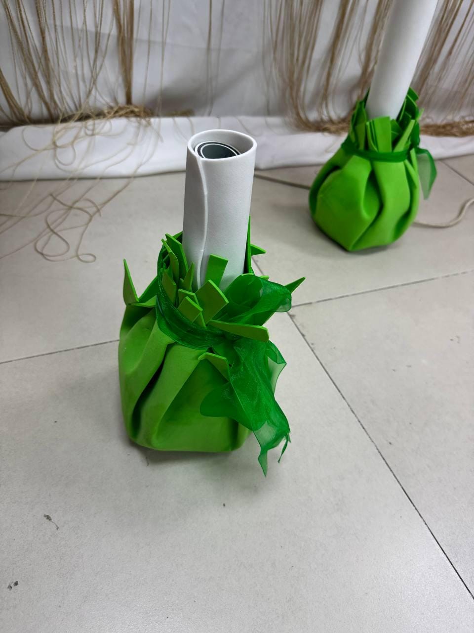 May include: Two decorative green fabric bases, each with a white cylindrical object rising from the center. The bases are wrapped in a pleated design, with green accents and a sheer green fabric. The objects are on a light gray surface.