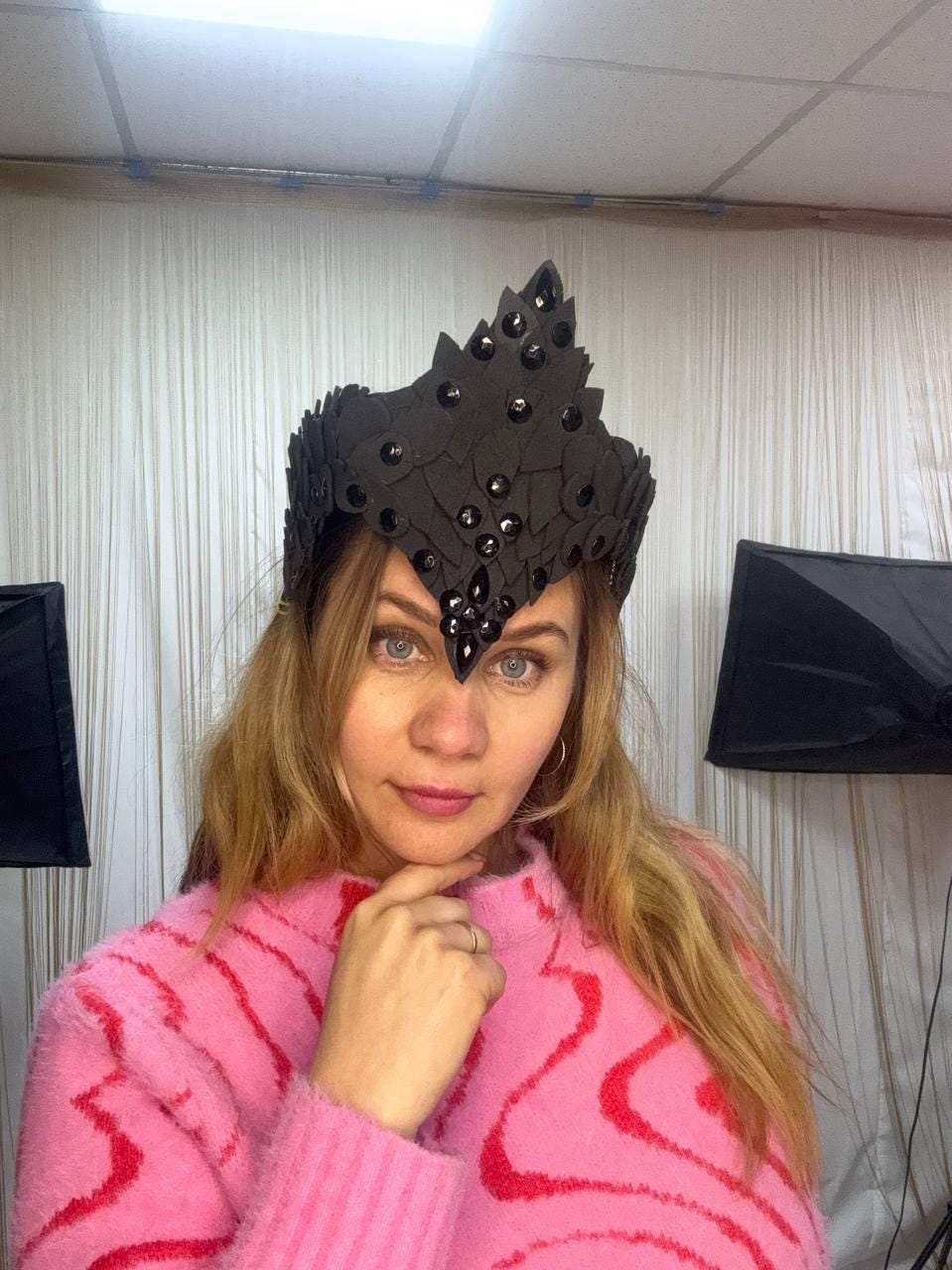 May include: A black crown with a leaf-like design and embedded dark gems. The crown is worn by a person wearing a pink sweater.
