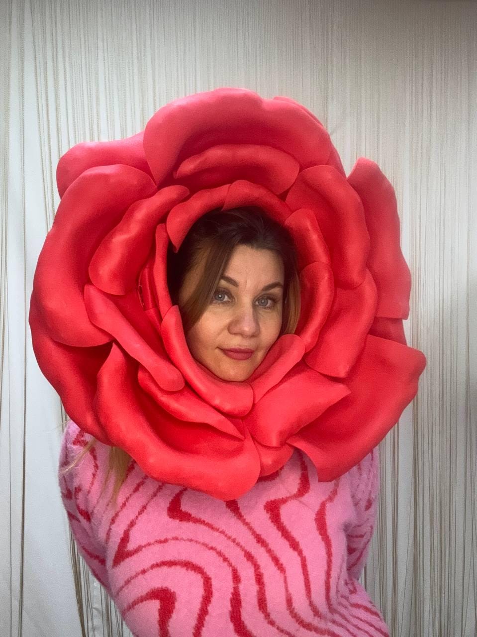 May include: A large, vibrant red rose costume frames a person's face. The rose is a bold, three-dimensional design. The person is wearing a pink sweater with a wavy pattern. The background is a neutral, textured wall.