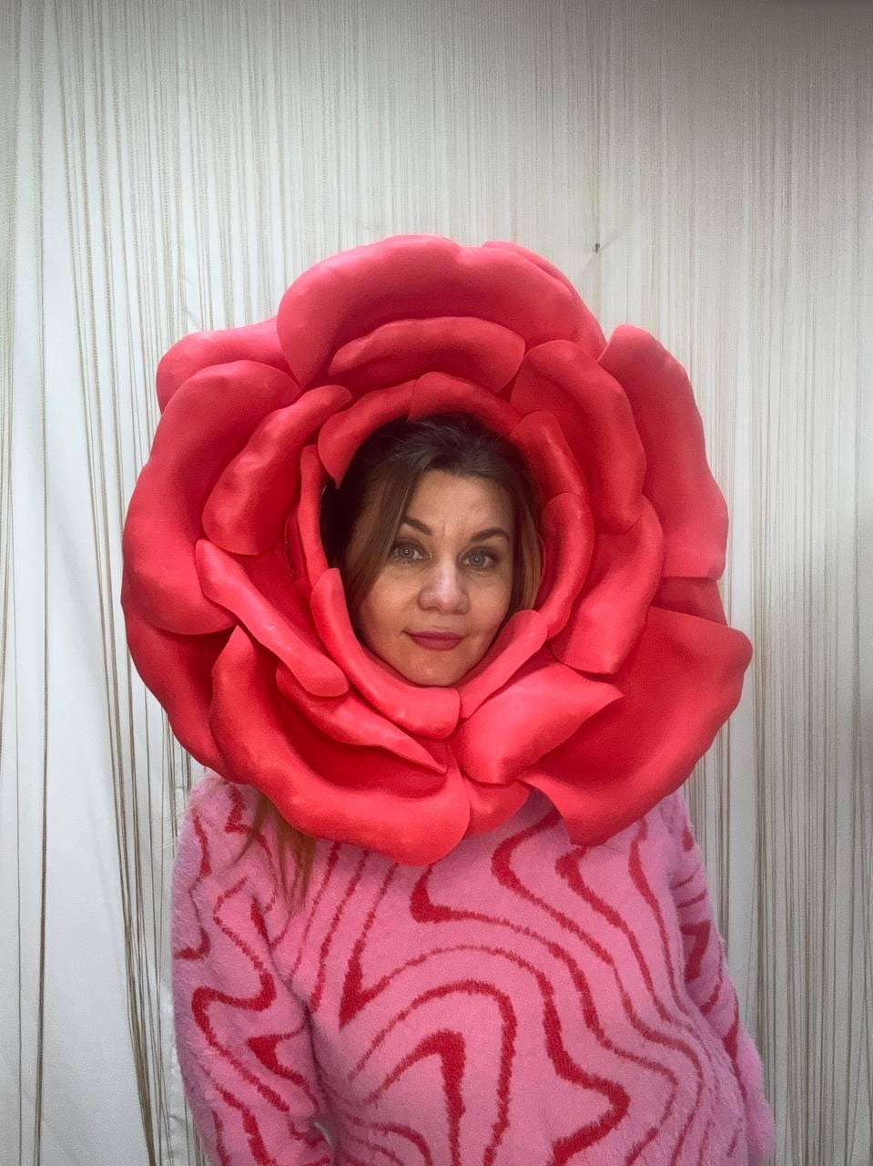 May include: A woman wearing a large, vibrant red rose headpiece. She is also wearing a pink sweater with a wavy, red pattern. The rose is the focal point, with its petals framing her face. The background is a neutral, vertical-striped curtain.