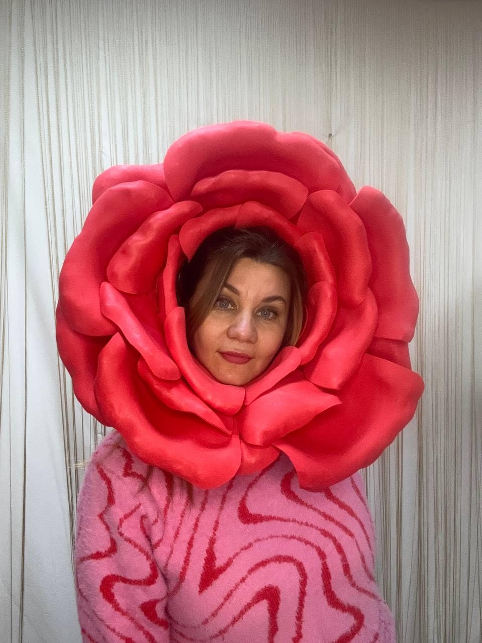 May include: A person wearing a large, vibrant red rose headpiece. The rose is the focal point, with detailed petals. The person is wearing a pink sweater with a wavy red pattern. The background is a white, textured surface.