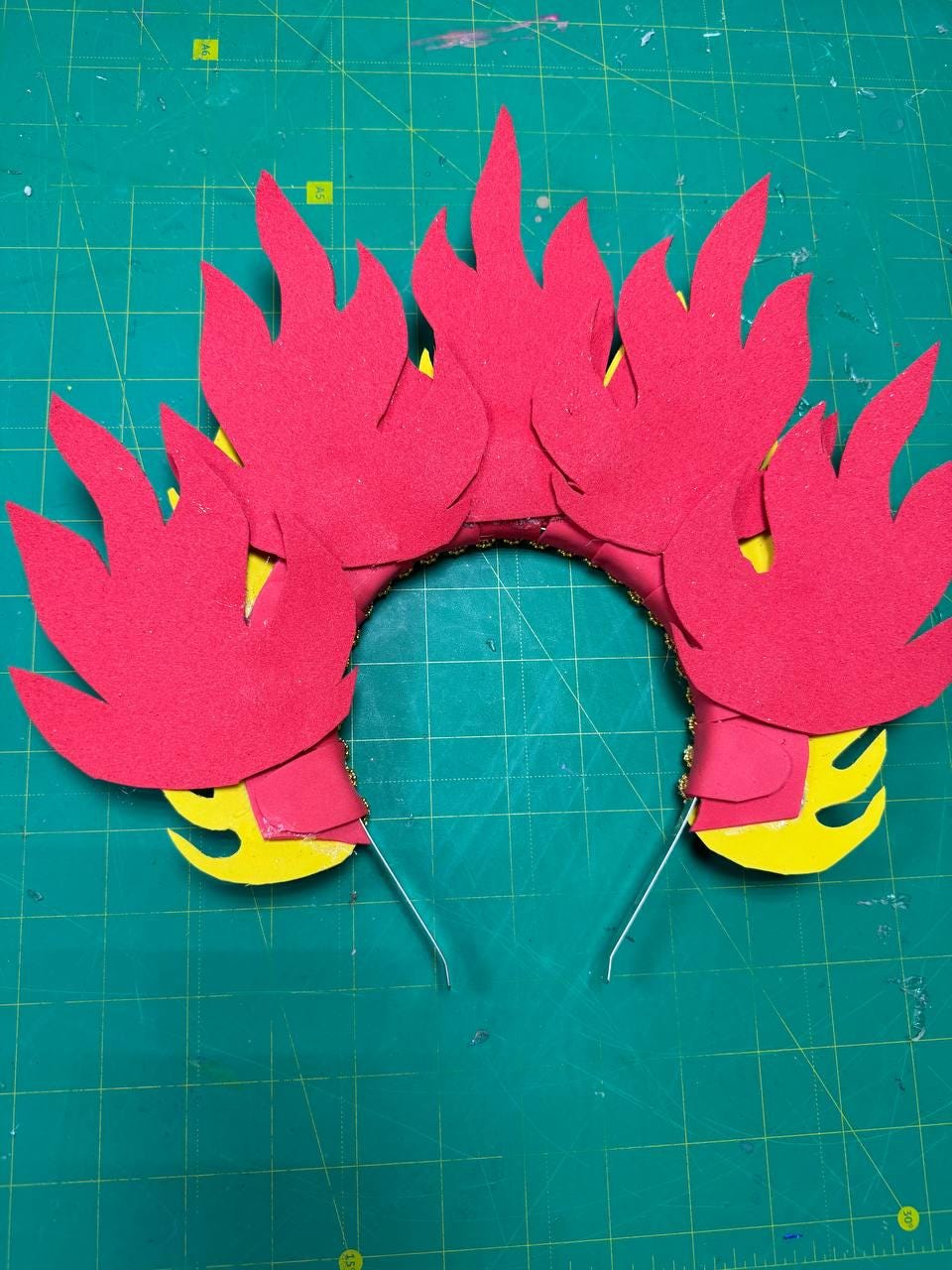 May include: A red felt flame crown with yellow accents. The crown features multiple flame shapes attached to a gold-colored headband. The flames are arranged around the headband, creating a fiery effect. The crown is designed for costume or cosplay use.