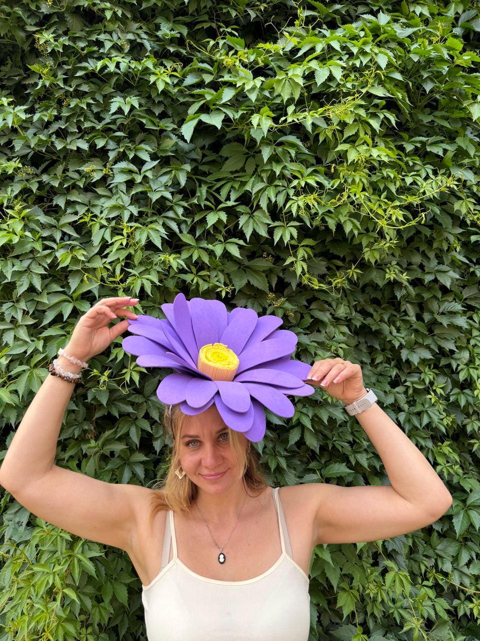 May include: A large, purple flower headpiece with a yellow center. The petals are a vibrant purple, and the center is a bright yellow. The headpiece is worn on the head. The background is a wall of green leaves.