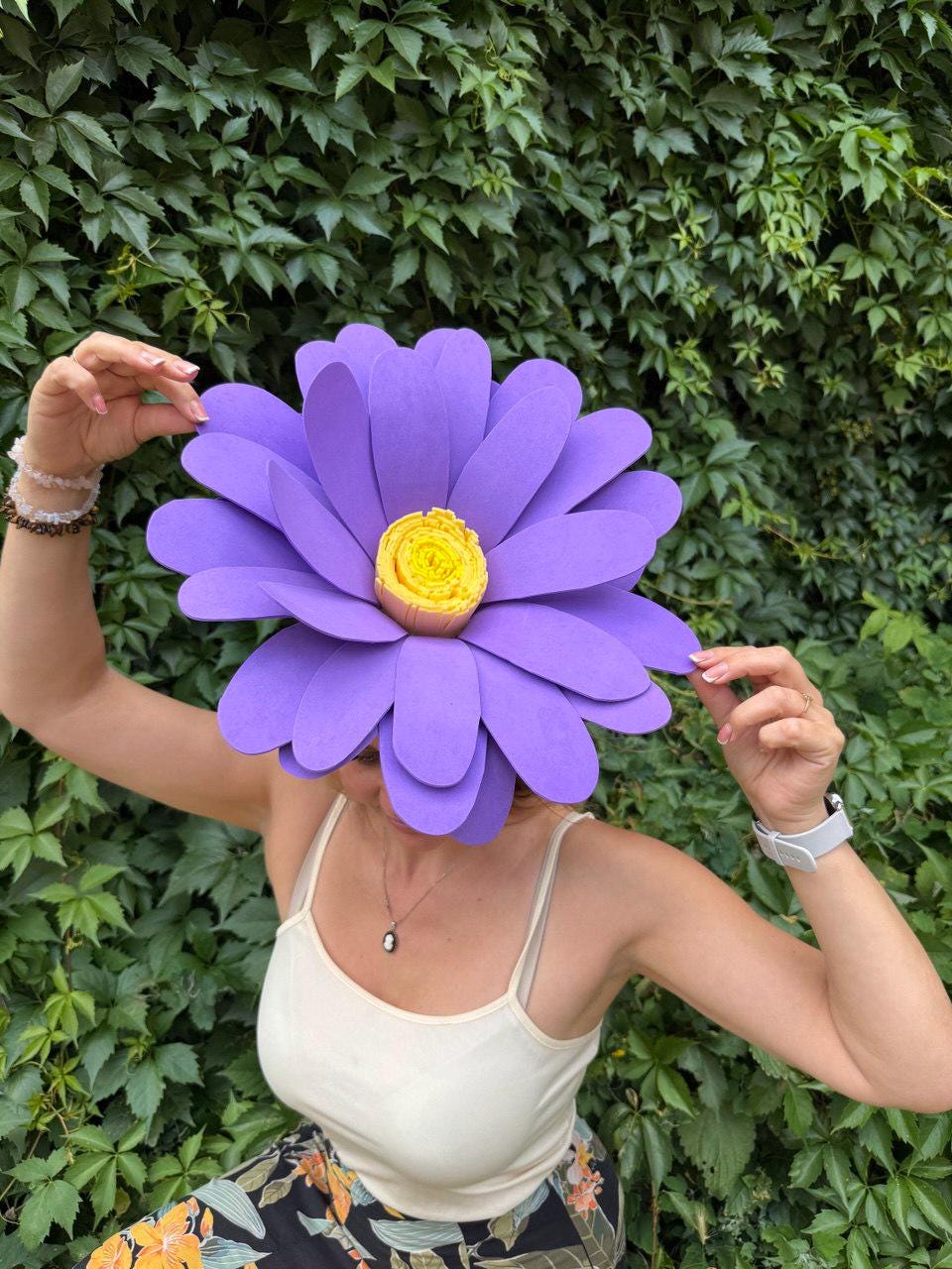 May include: A large, purple paper flower with a yellow center. The flower is held up in front of a person's face, obscuring it. The background is a wall of green leaves.