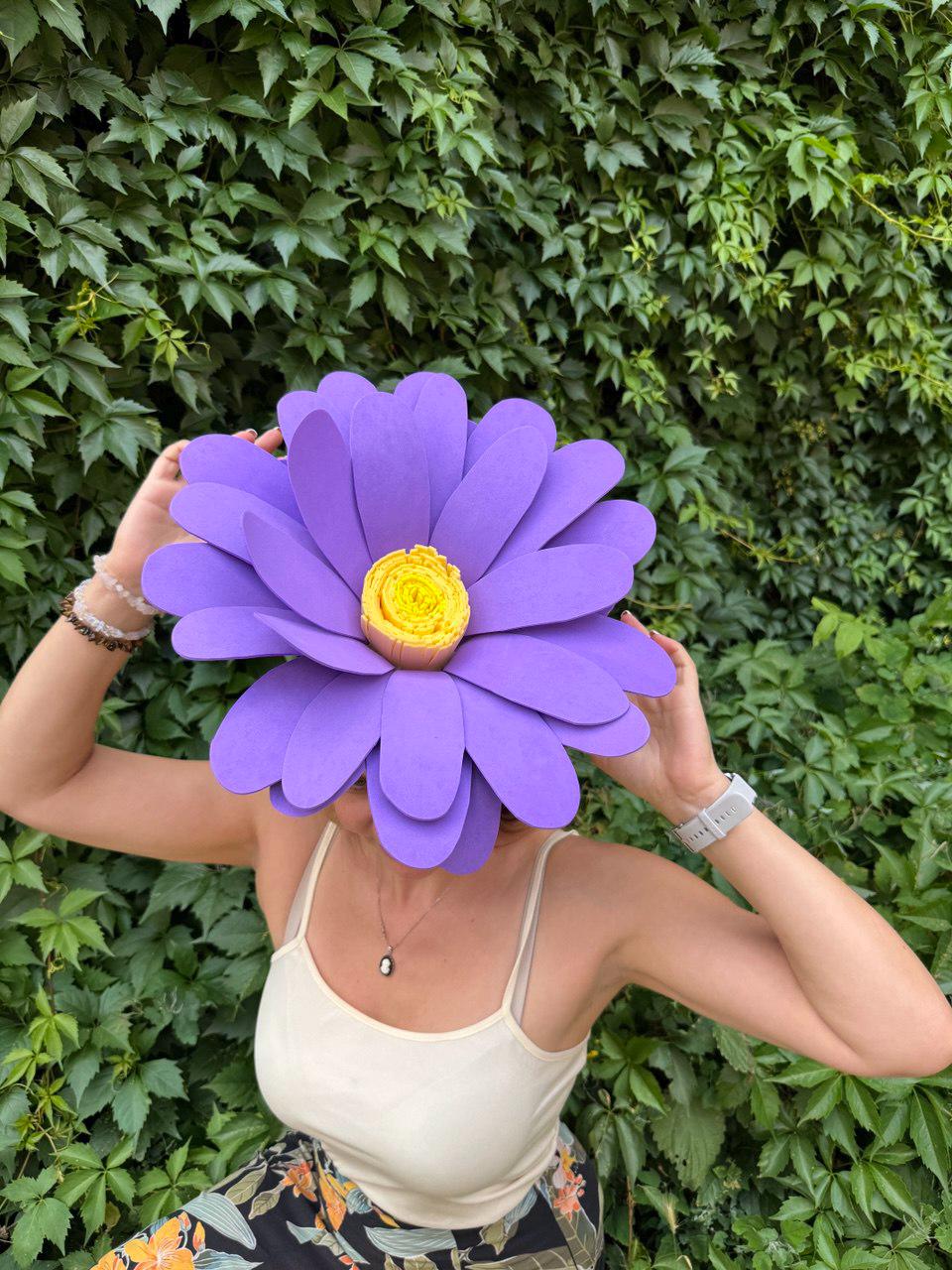 May include: A large, purple paper flower with a yellow center is held up in front of a person's face. The flower has multiple layers of petals. The background is a wall of green leaves.