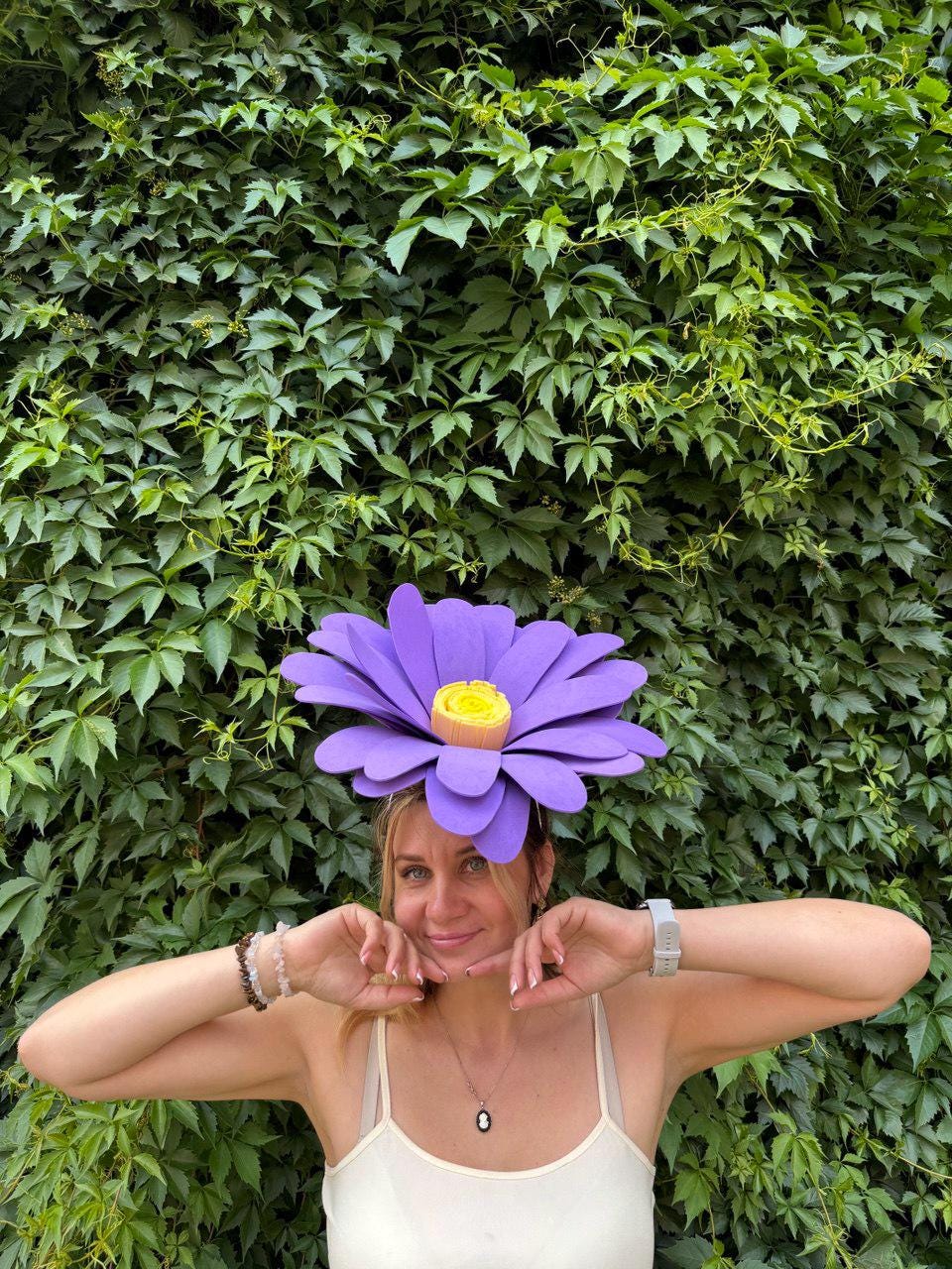 May include: A large, purple flower headpiece with a yellow center. The flower is worn on the head. The background is a wall of green leaves.