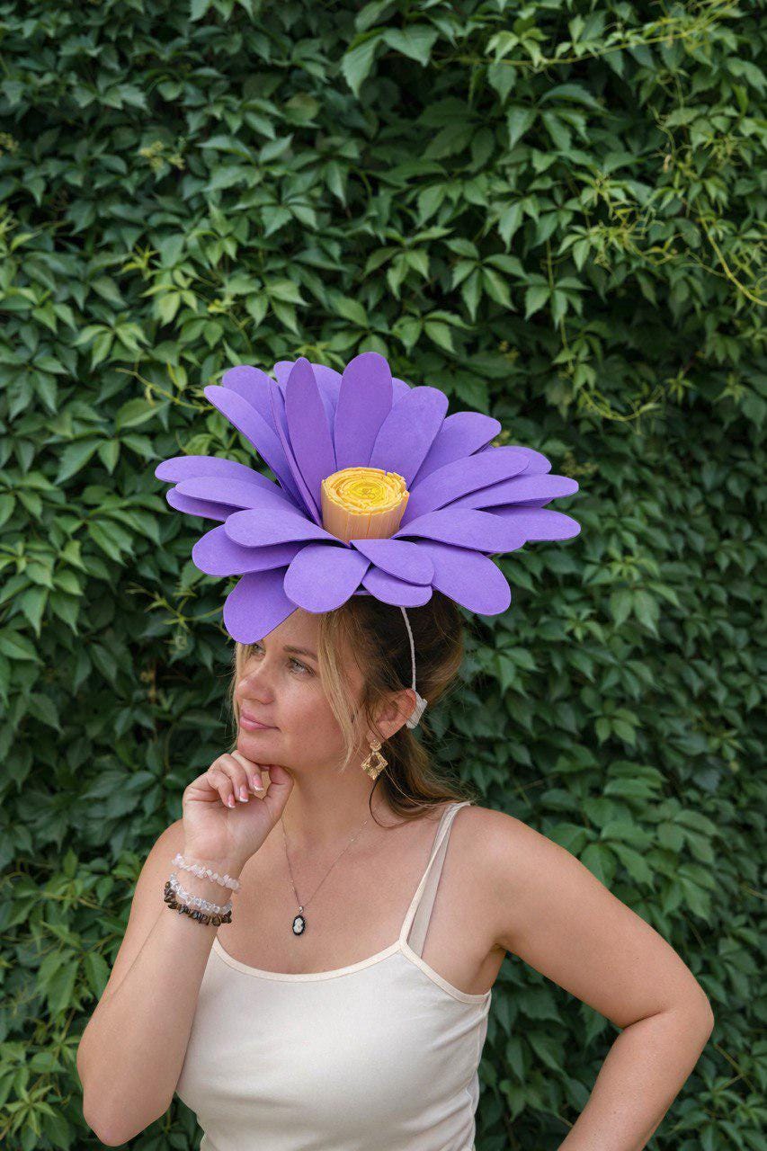 May include: A large, purple flower headpiece with a yellow center. The petals are layered and create a dramatic effect. The headpiece is worn on the head with a thin, white band. The background is a lush green wall of leaves.