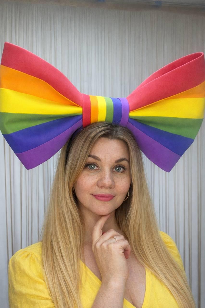 May include: A large, colorful rainbow bow headband. The bow features the colors of the rainbow: red, orange, yellow, green, blue, and purple. The headband is worn by a person with blonde hair and a yellow top.