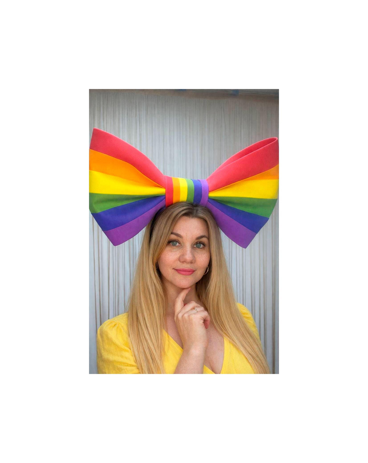 May include: A large rainbow-colored bow headband. The bow features the colors of the rainbow: red, orange, yellow, green, blue, and purple. The headband is worn by a person with long blonde hair and a yellow top.