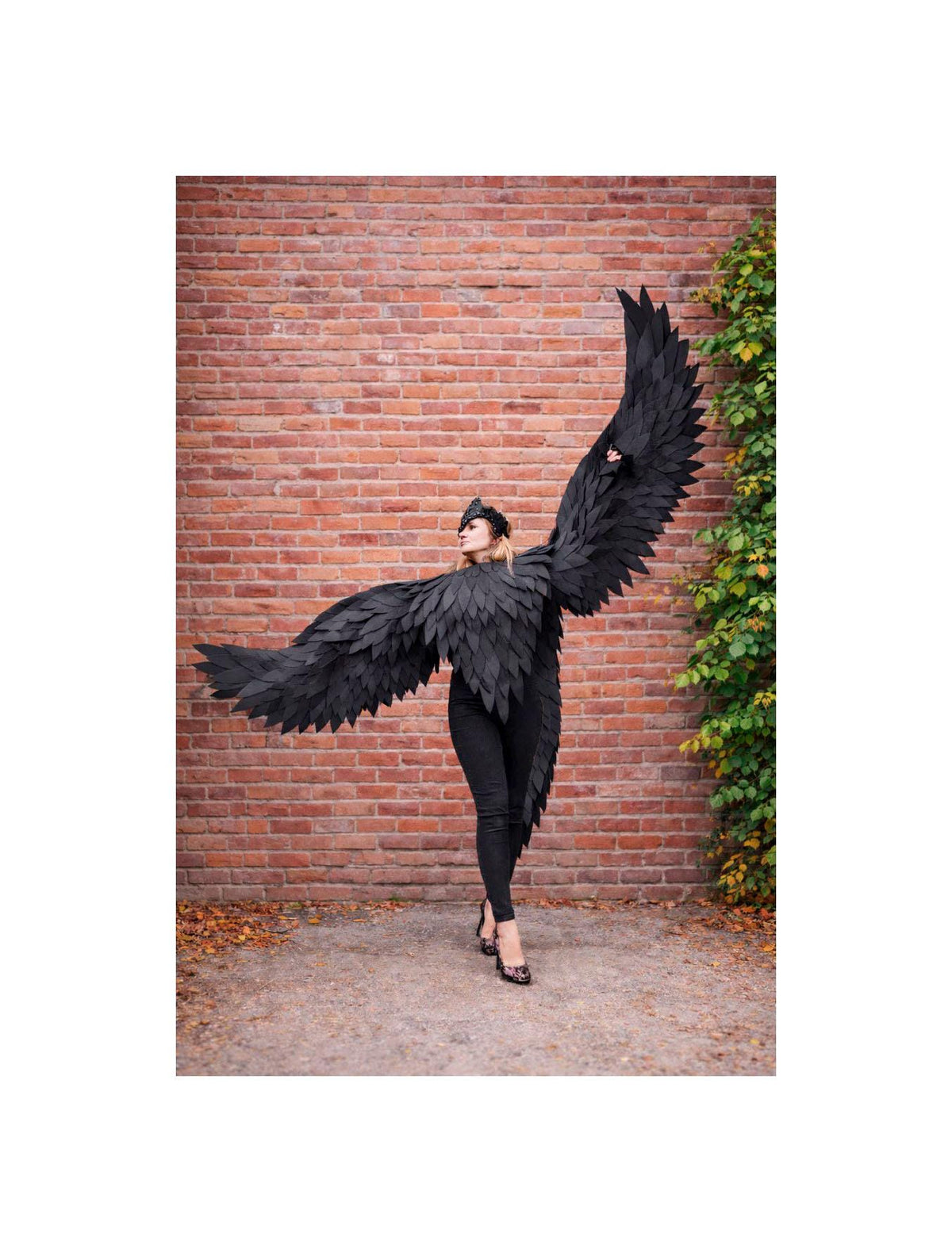May include: A person wearing a black feathered wing costume with a matching headpiece, posing in front of a brick wall. The wings are large and spread out, creating a dramatic effect. The outfit is all black, including the pants and shoes.