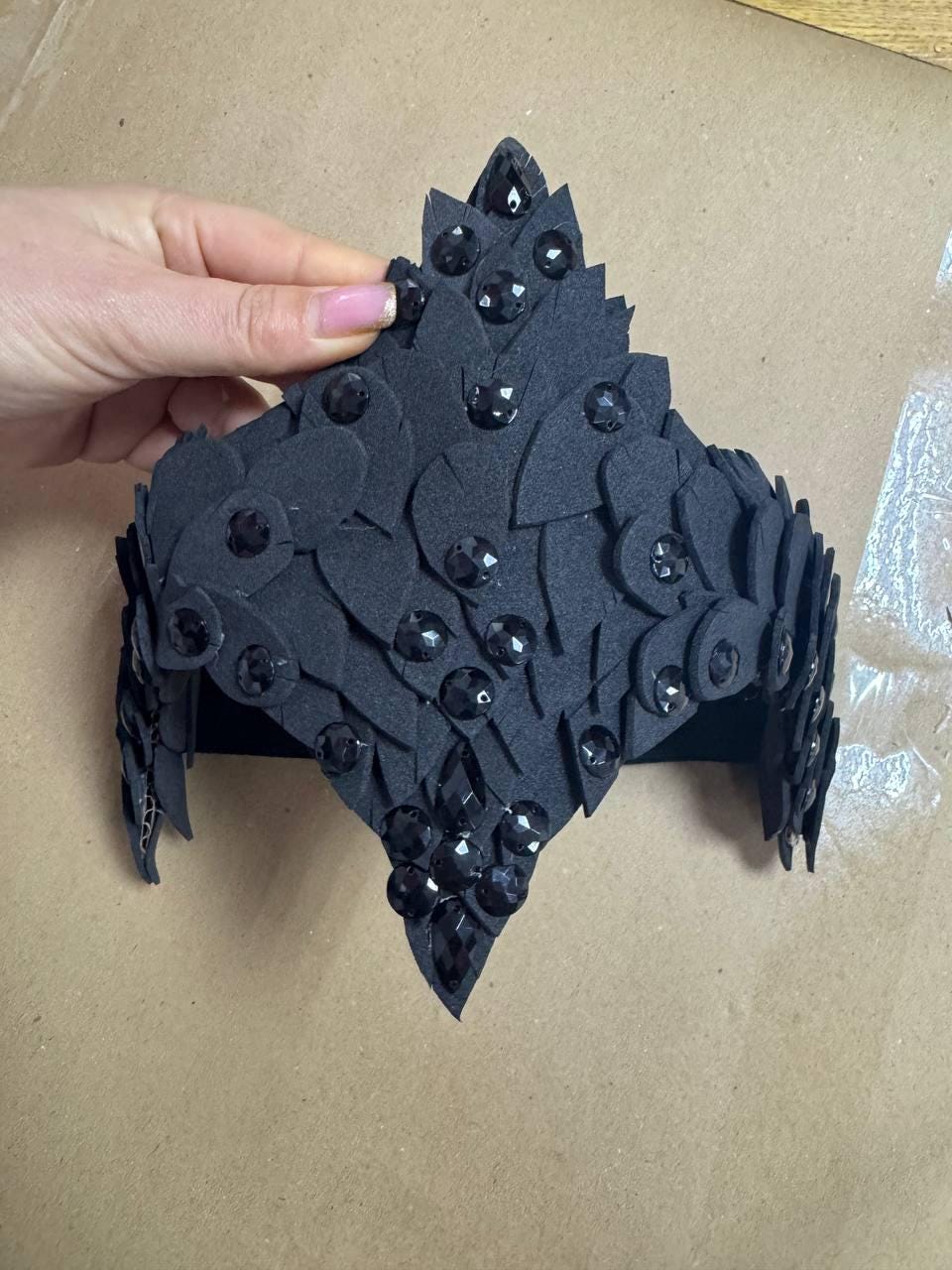 May include: A black crown-shaped headpiece with a leaf-like design, adorned with numerous black gemstones. The headpiece is made of a dark, textured material and is held by a hand. The crown is designed to be worn on the head.