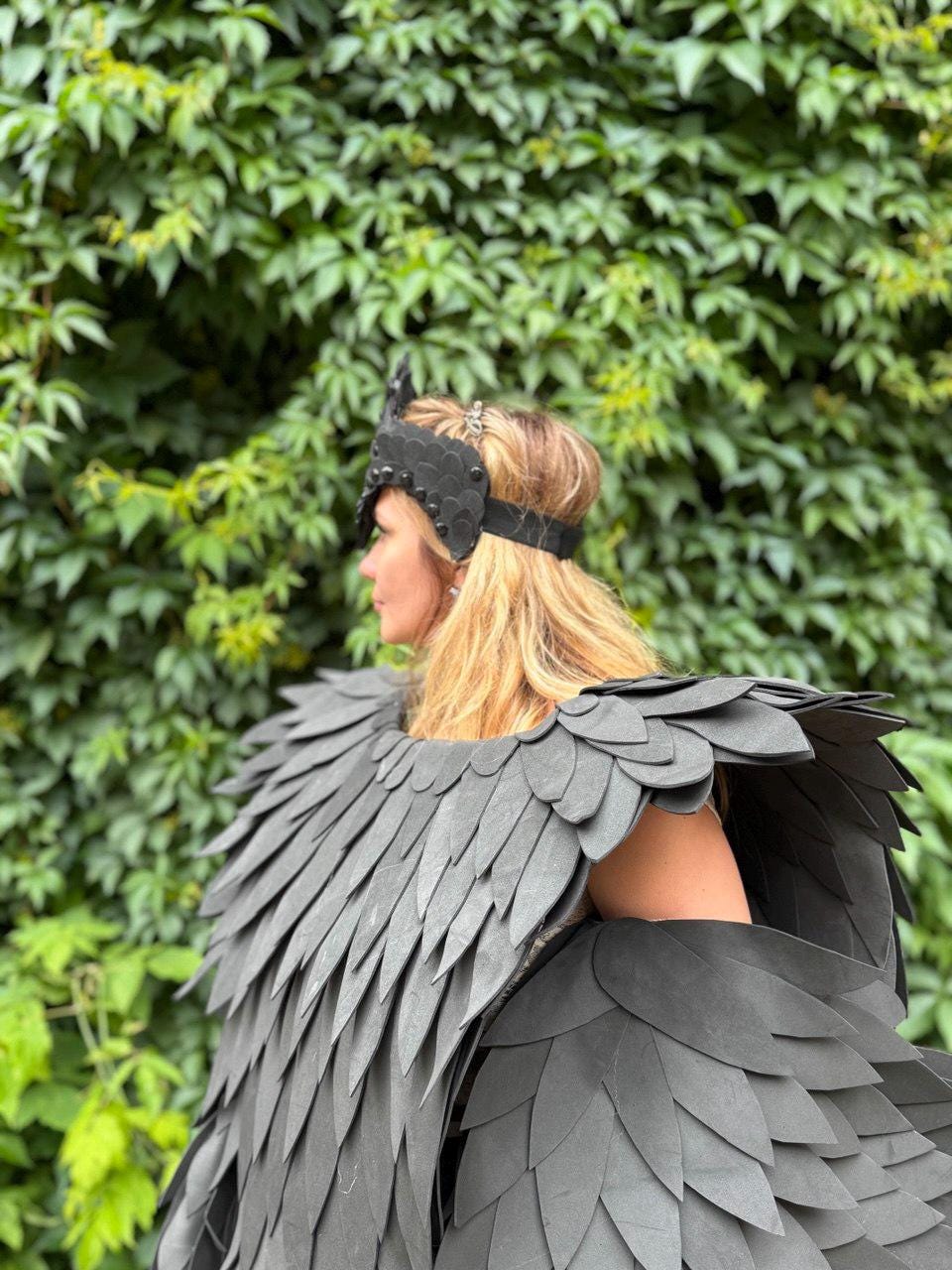 May include: A black swan costume featuring a feathered cape and headpiece. The cape is made of layered, dark gray feathers, and the headpiece has a matching design. The costume is worn against a backdrop of green foliage.