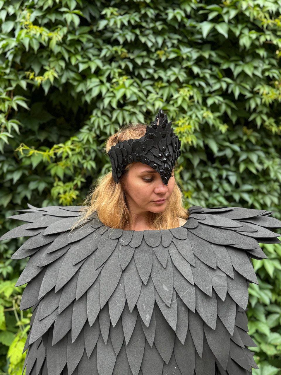 May include: A black costume featuring a feathered shoulder piece and a matching crown. The crown is adorned with dark embellishments. The costume is worn against a backdrop of green foliage, creating a striking contrast.