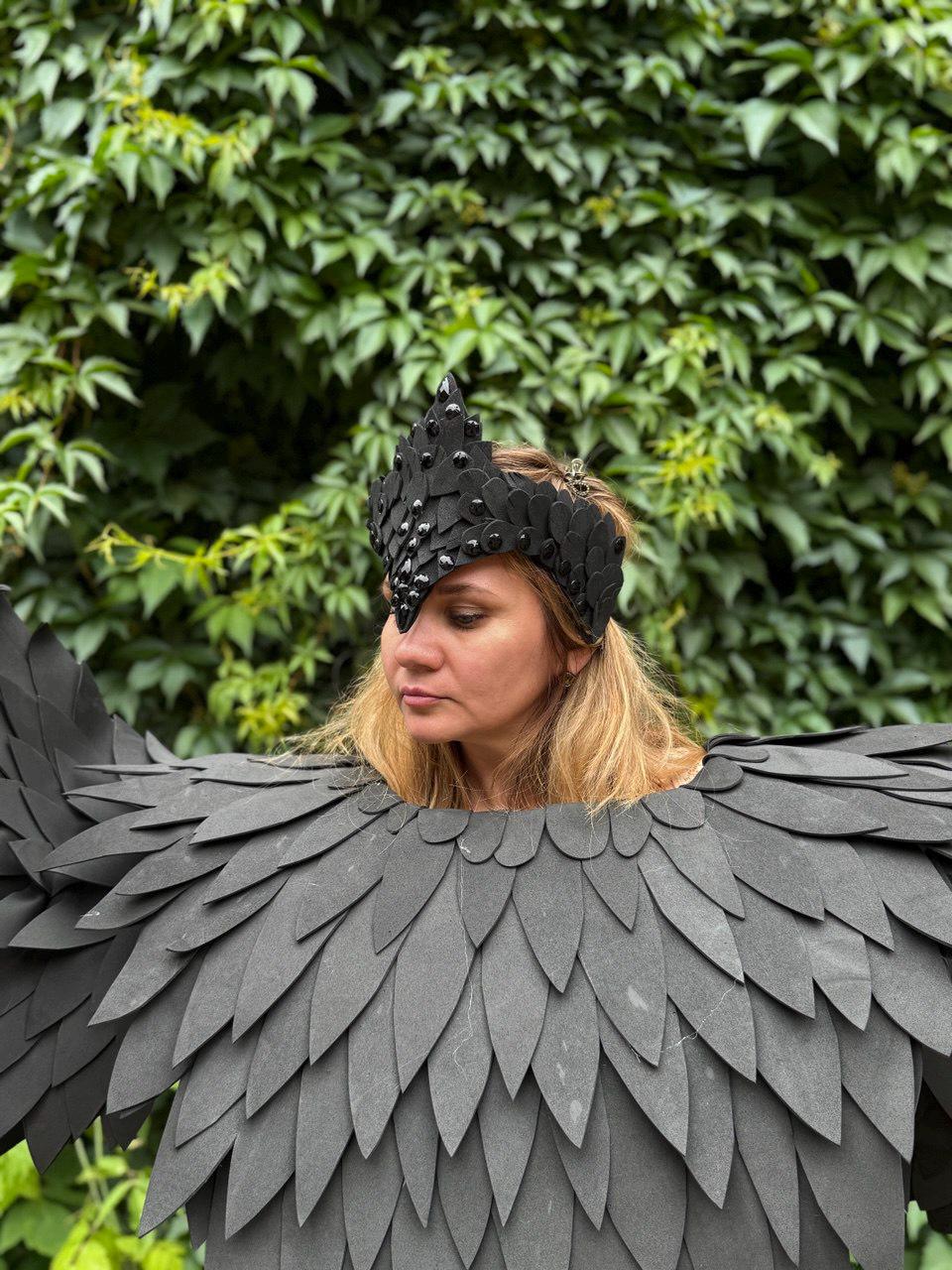 May include: A black costume featuring a feathered wing design and a matching crown. The wings are made of layered, dark gray, leaf-shaped pieces. The crown is black with small, dark embellishments. The background is a lush green foliage.