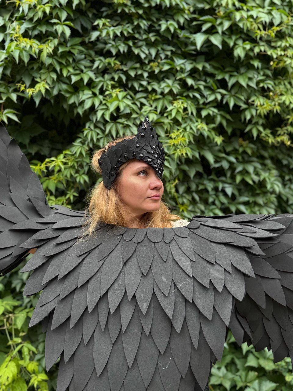 May include: A black costume with large, layered wings and a matching headpiece. The wings are made of individual, dark gray, feather-shaped pieces. The headpiece is adorned with small, dark studs. The background is a wall of green leaves.