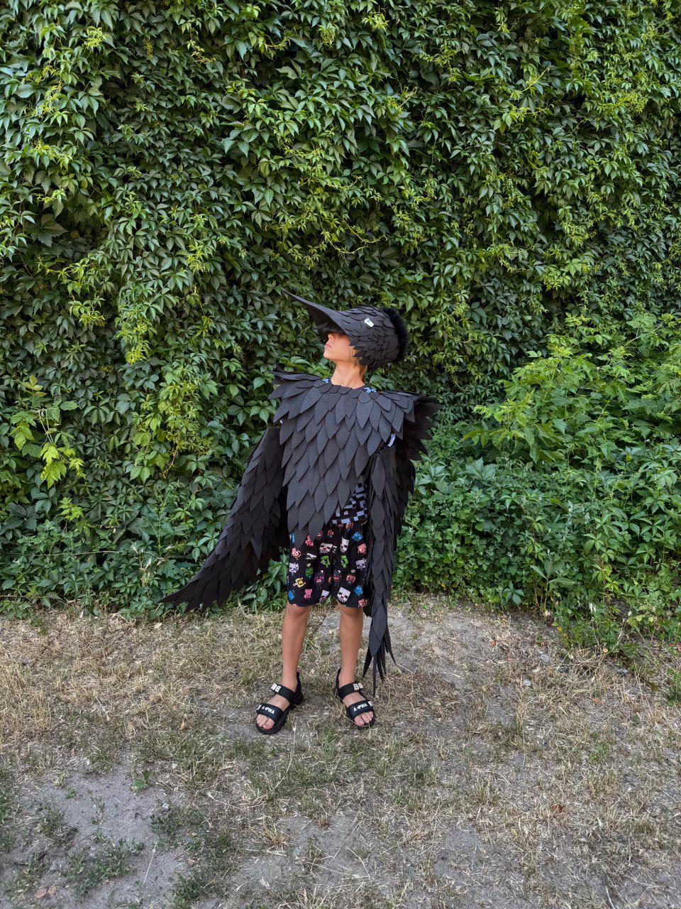 May include: A person wearing a black bird costume with a long beak, feathered wings, and a matching headpiece. The costume is made of dark material and the person is wearing black sandals. The background is a wall of green foliage.