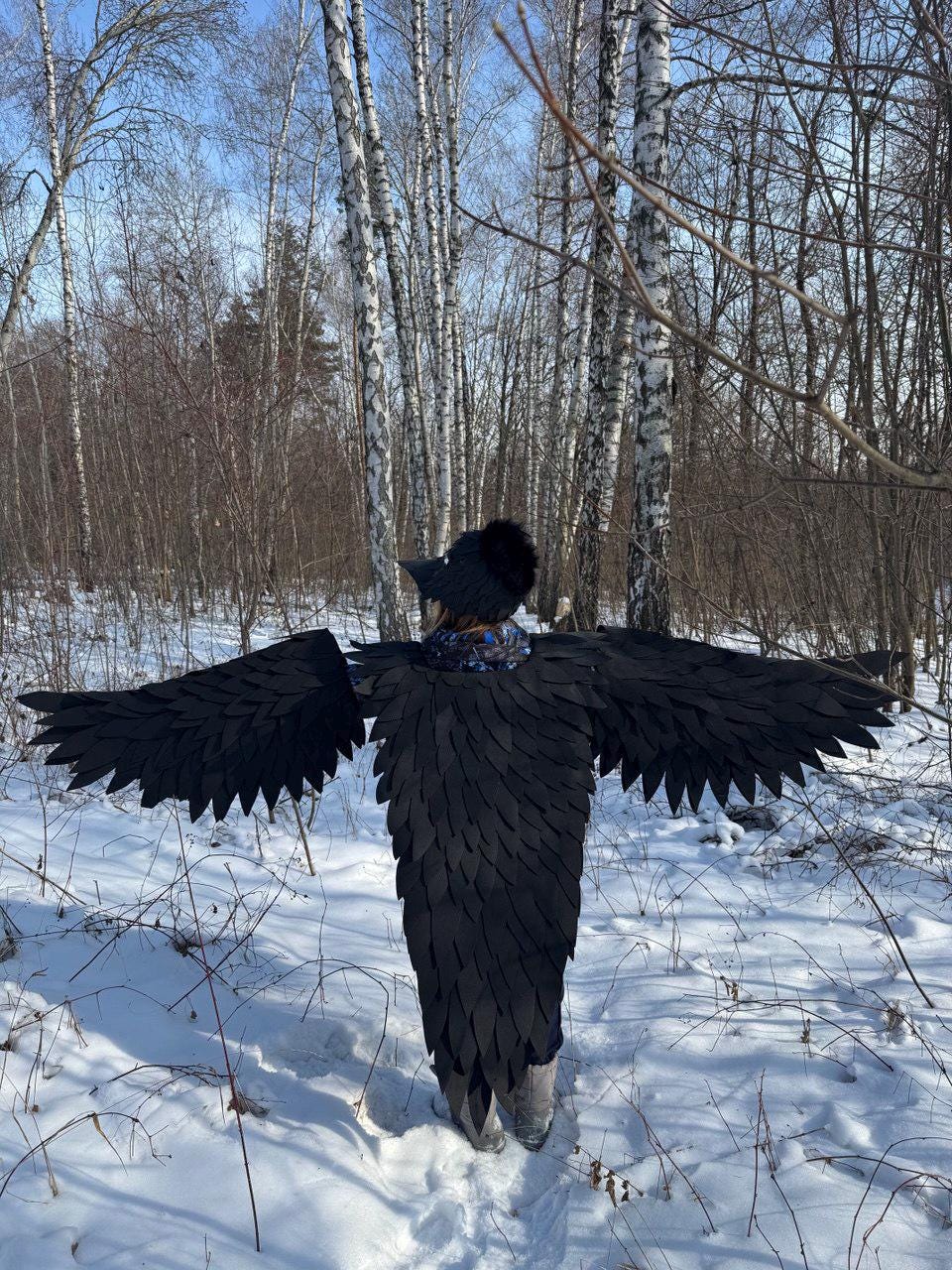 May include: A black costume with large, feathered wings, worn in a snowy forest setting. The wings are spread wide, and the costume covers the body. The background features bare trees and snow on the ground.
