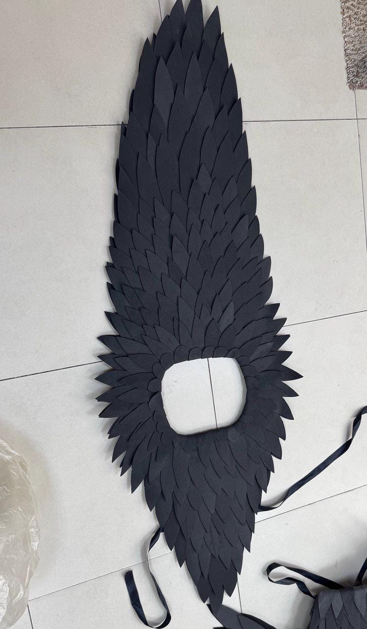 May include: A large, black costume piece resembling a feather-covered wing. The wing has a central opening and is made of layered, dark-colored paper or cardstock. Black ribbons are attached for securing the costume.