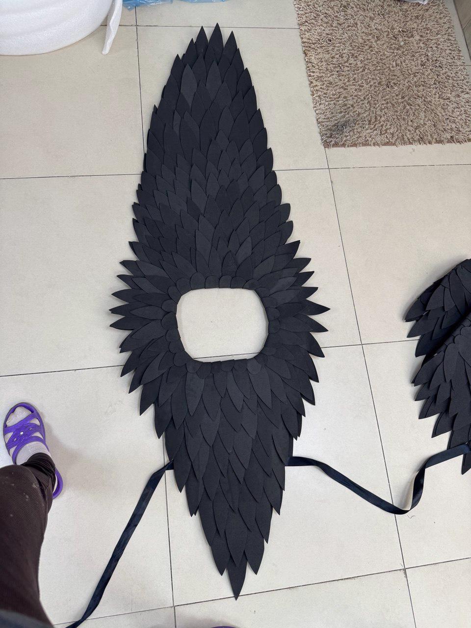 May include: Black costume wings with a central opening, constructed from layered, leaf-shaped pieces. A black ribbon is attached. The wings are designed for a costume or theatrical use.
