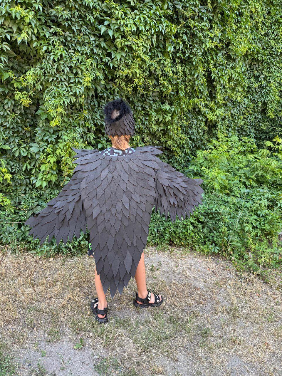 May include: A black raven costume with layered feather details, including a matching headpiece. The costume has large, spread wings and is worn by a person standing outdoors. The background features a wall of green foliage and dry grass.