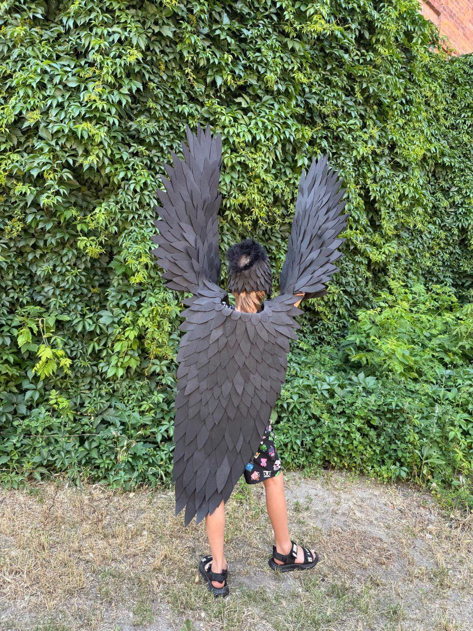 May include: A child wearing a black raven costume with large, layered wings and a matching headpiece. The costume is made of dark gray material and is set against a backdrop of green foliage and grass.