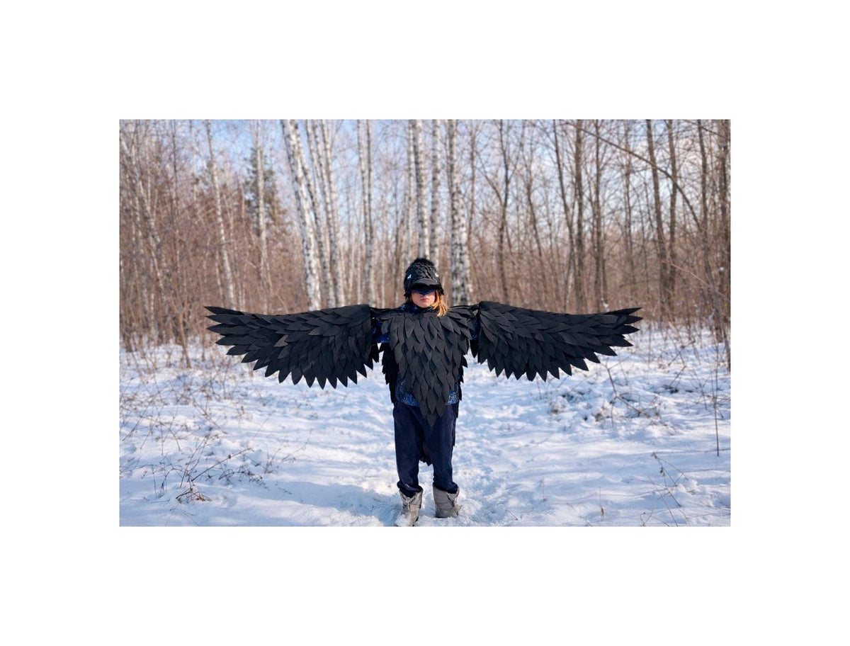 May include: A child wearing a black raven costume with large, spread wings. The costume includes a feathered vest and a headpiece. The background is a snowy forest with bare trees.