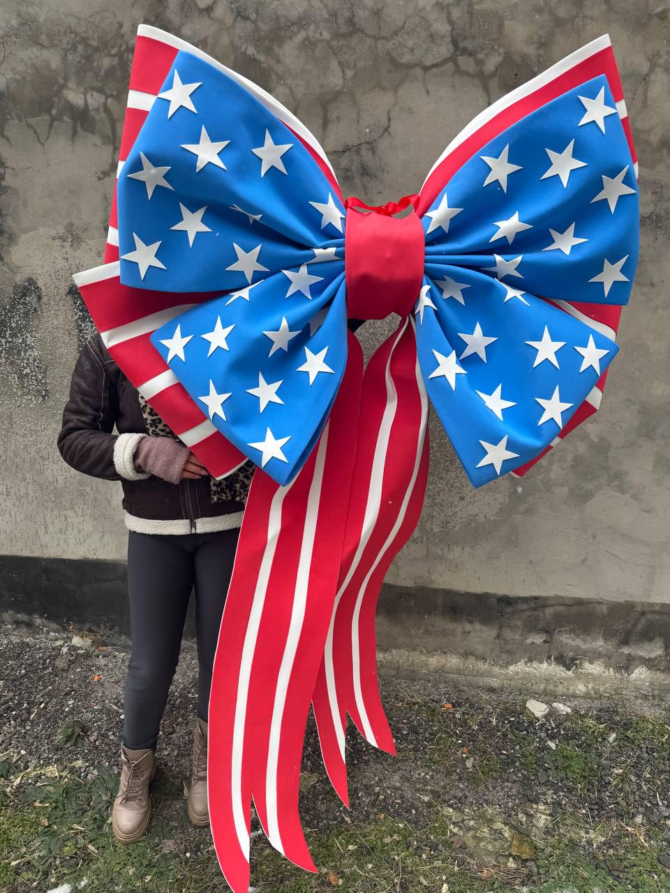 May include: A large decorative bow featuring a patriotic design. The bow has a blue center adorned with white stars, red and white striped ribbons, and a red center knot. This festive bow is suitable for various celebratory occasions.