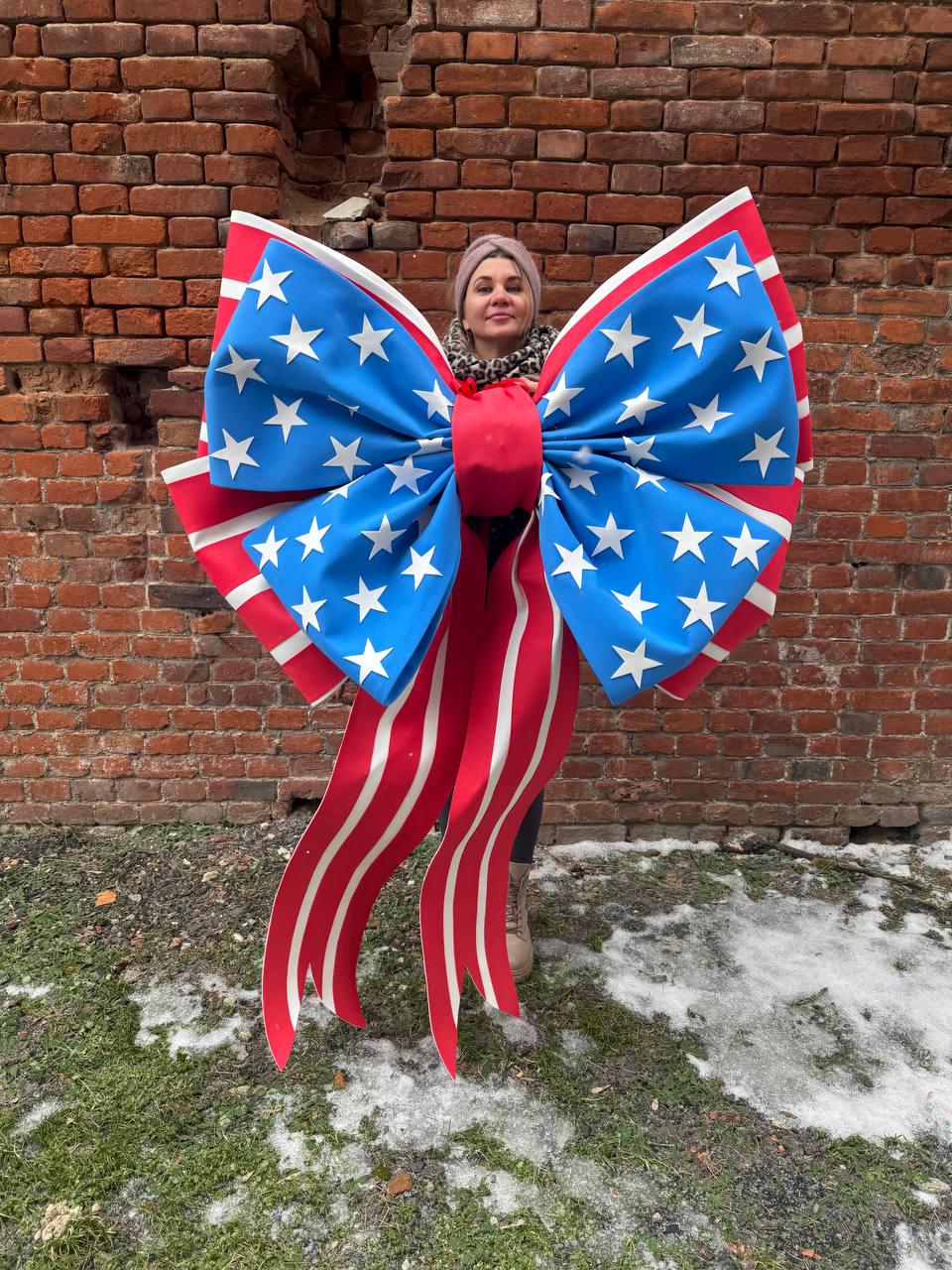 May include: A large decorative bow featuring the American flag design. The bow has a blue center with white stars, red and white striped edges, and long red and white striped streamers. The bow is held in front of a brick wall.