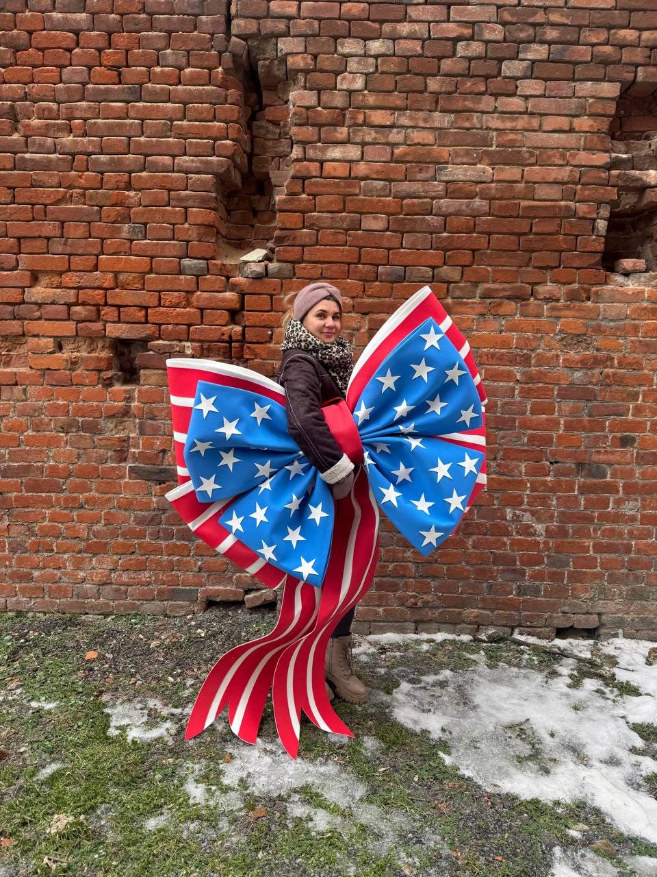 May include: A large bow featuring the American flag design. The bow has a blue center with white stars and red and white striped ribbons. The bow is held in front of a brick wall.