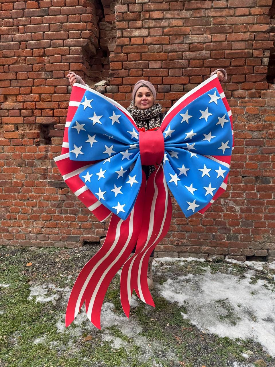May include: A large decorative bow featuring the American flag design. The bow has a blue center with white stars, red and white striped ribbons, and a red center knot. The bow is held up in front of a brick wall.