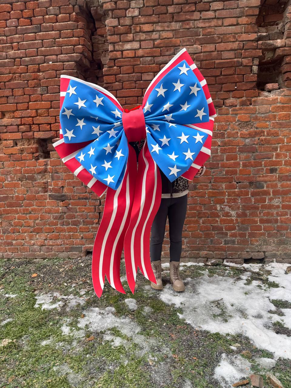 May include: A large decorative bow with a patriotic design. The bow features a blue center adorned with white stars, red and white striped edges, and long red and white striped ribbons. The bow is held in front of a brick wall.
