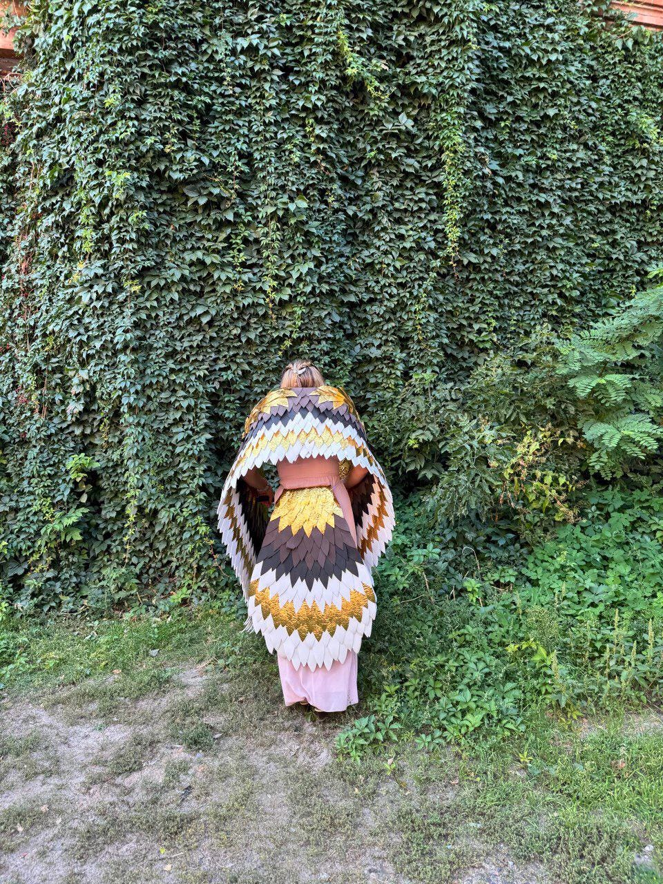 May include: A person wearing a layered, feathered cape with a color scheme of white, gold, brown, and black. The cape is worn over a pink dress. The background features a wall covered in green ivy and lush greenery.
