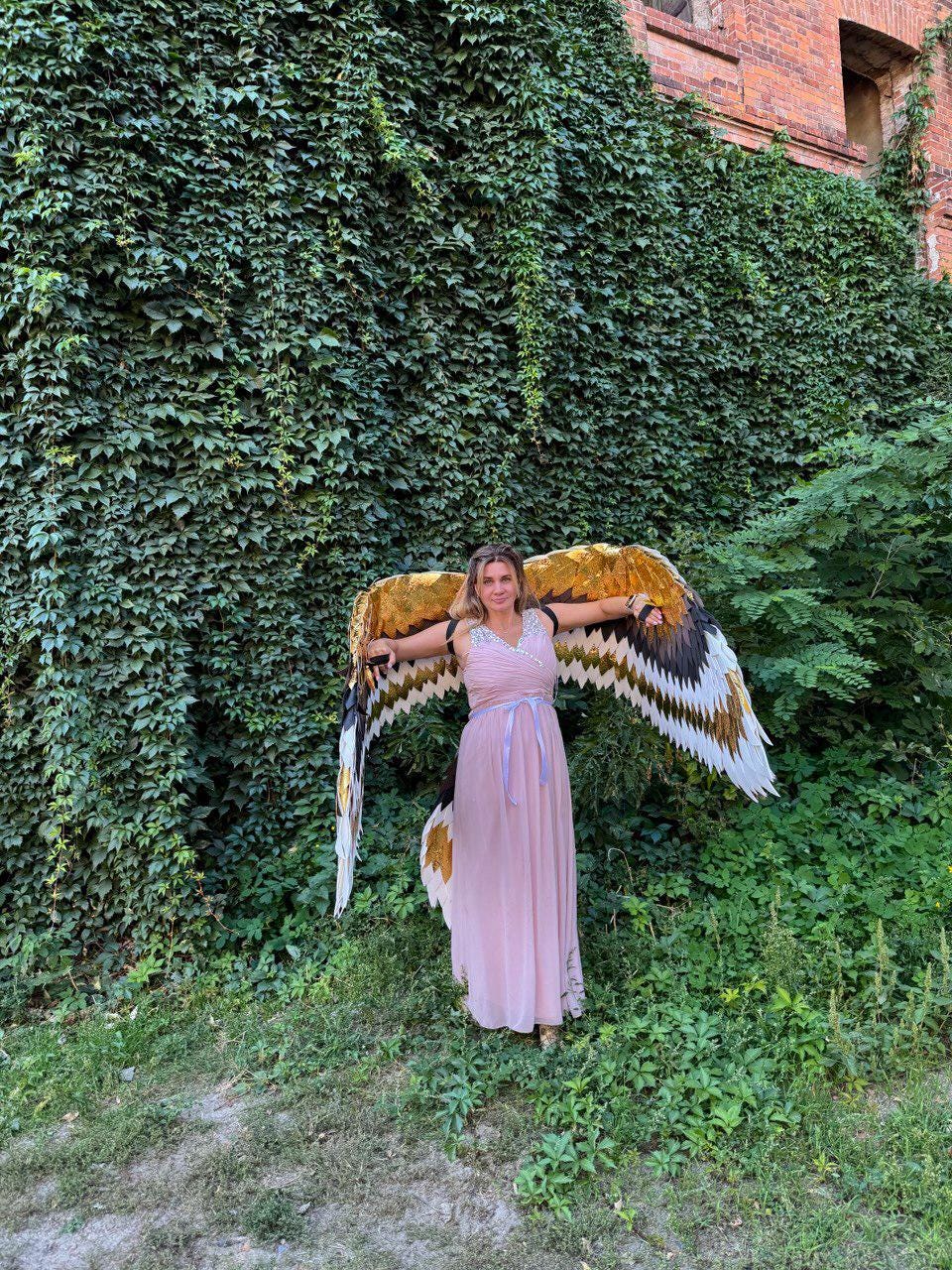 May include: A person wearing a long, light pink dress and large, decorative bird wings with gold, black, and white feathers. The wings are spread wide against a backdrop of green foliage and a brick building.