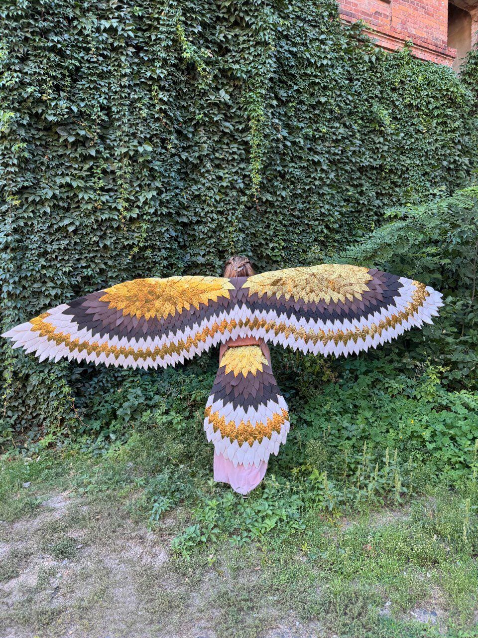 May include: A person wearing a large, handmade bird wing costume. The wings feature a layered design with shades of brown, gold, and white, resembling feathers. The costume is set against a backdrop of lush green foliage.
