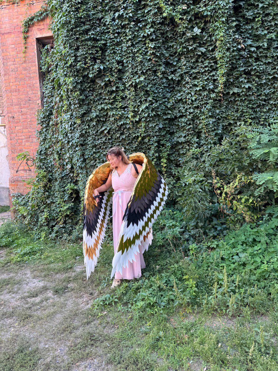 May include: A woman wearing large, feathered wings with gold, brown, black, and white sections. The wings are spread wide, and the woman is wearing a pink dress. The background features a brick building covered in green vines and lush greenery.