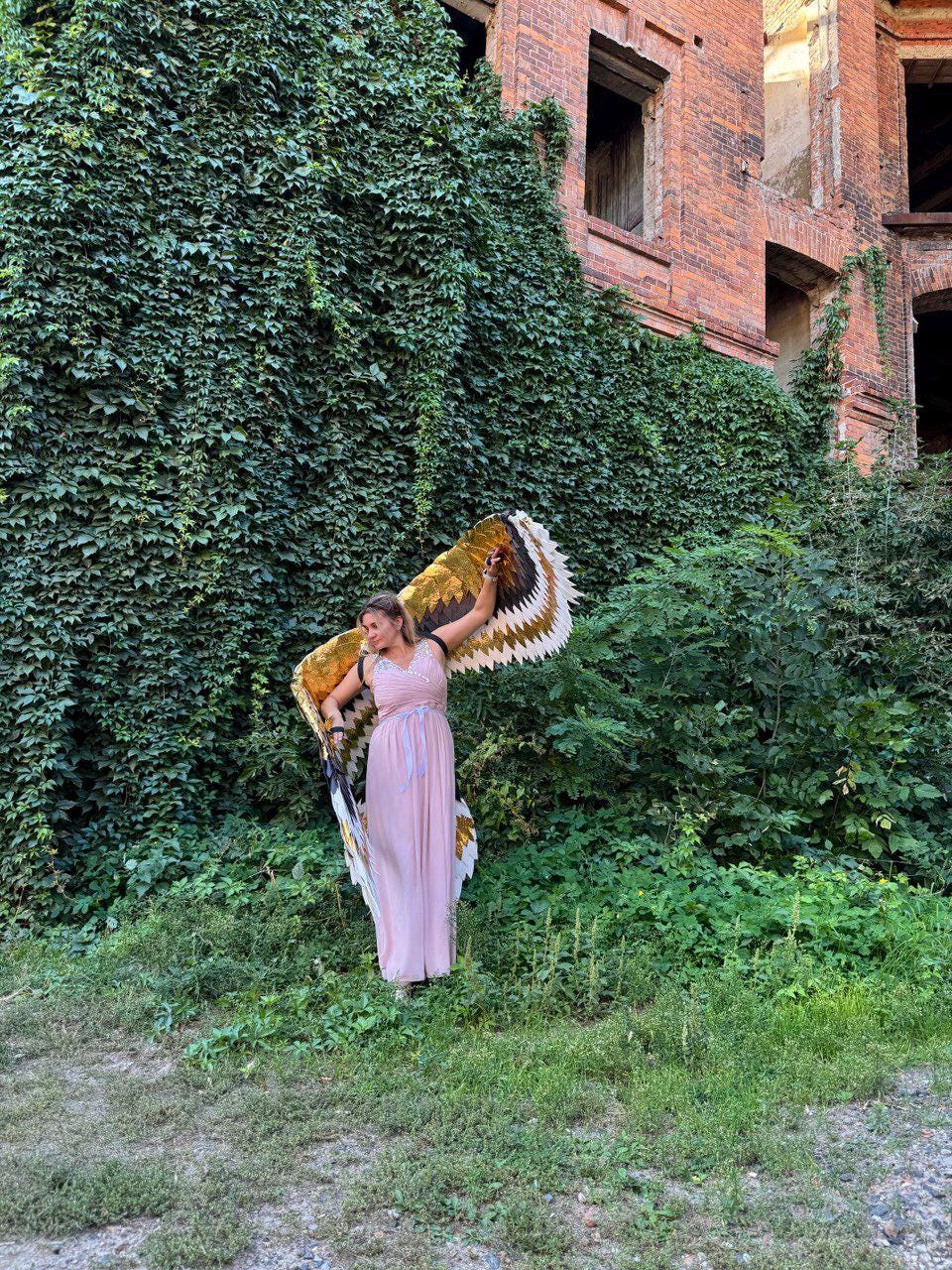 May include: A person wearing a long, light pink dress and large, decorative wings with brown, gold, and white feather patterns. The person is standing in front of a wall covered in green vines and a brick building.