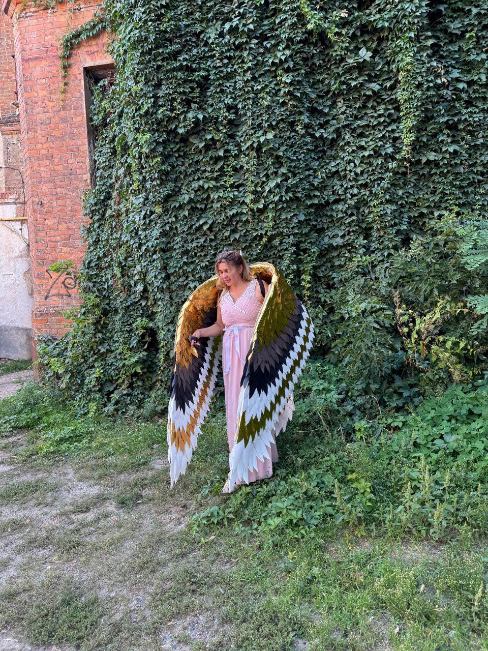 May include: A person wearing large, colorful angel wings with a pink dress. The wings feature a layered design with gold, brown, black, and white feathers. The background includes a brick building covered in green vines and lush greenery.