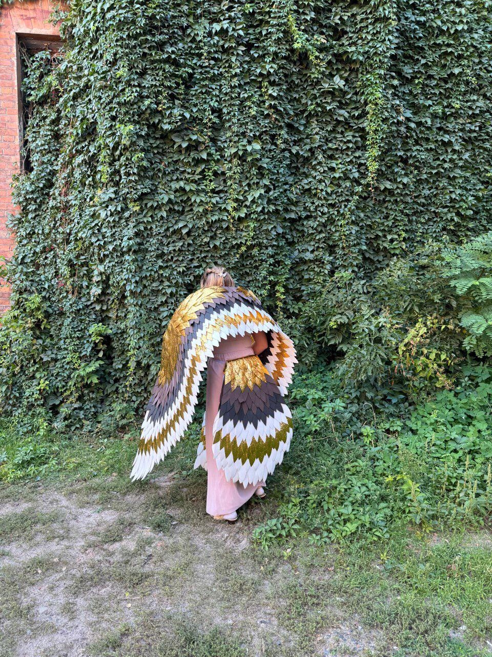 May include: A person wearing a large, colorful wing costume with gold, brown, white, and green feather-like details. The costume is worn in front of a wall of green foliage and grass.