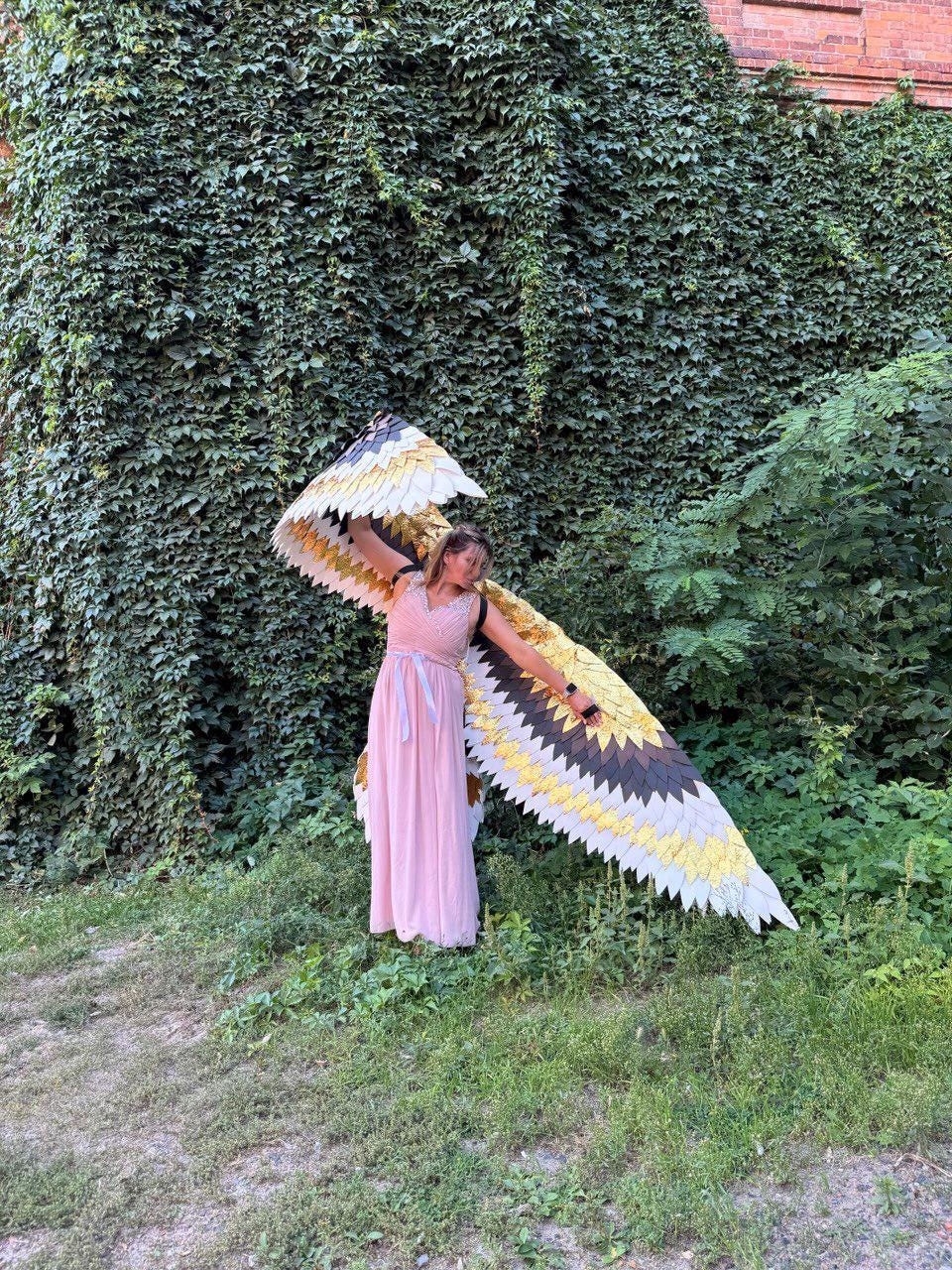 May include: A person wearing large, decorative bird wings with a color scheme of white, gold, black, and yellow. The wings are spread wide, and the person is wearing a pink dress. The background features lush green foliage.