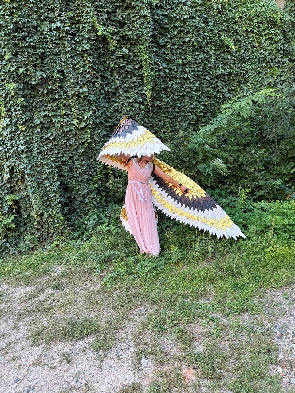 May include: A person wearing a pink dress and large, decorative bird wings. The wings feature a pattern of black, white, and yellow feathers. The background includes a wall of green foliage and grassy ground.