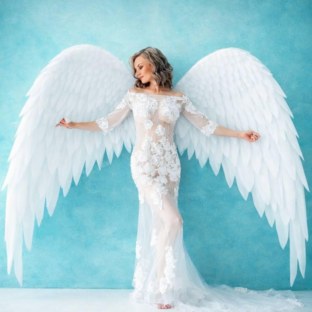 May include: A woman wearing a white, floor-length gown with lace detailing and large white feathered angel wings. The background is a light blue. The gown has long sleeves and a sheer skirt.